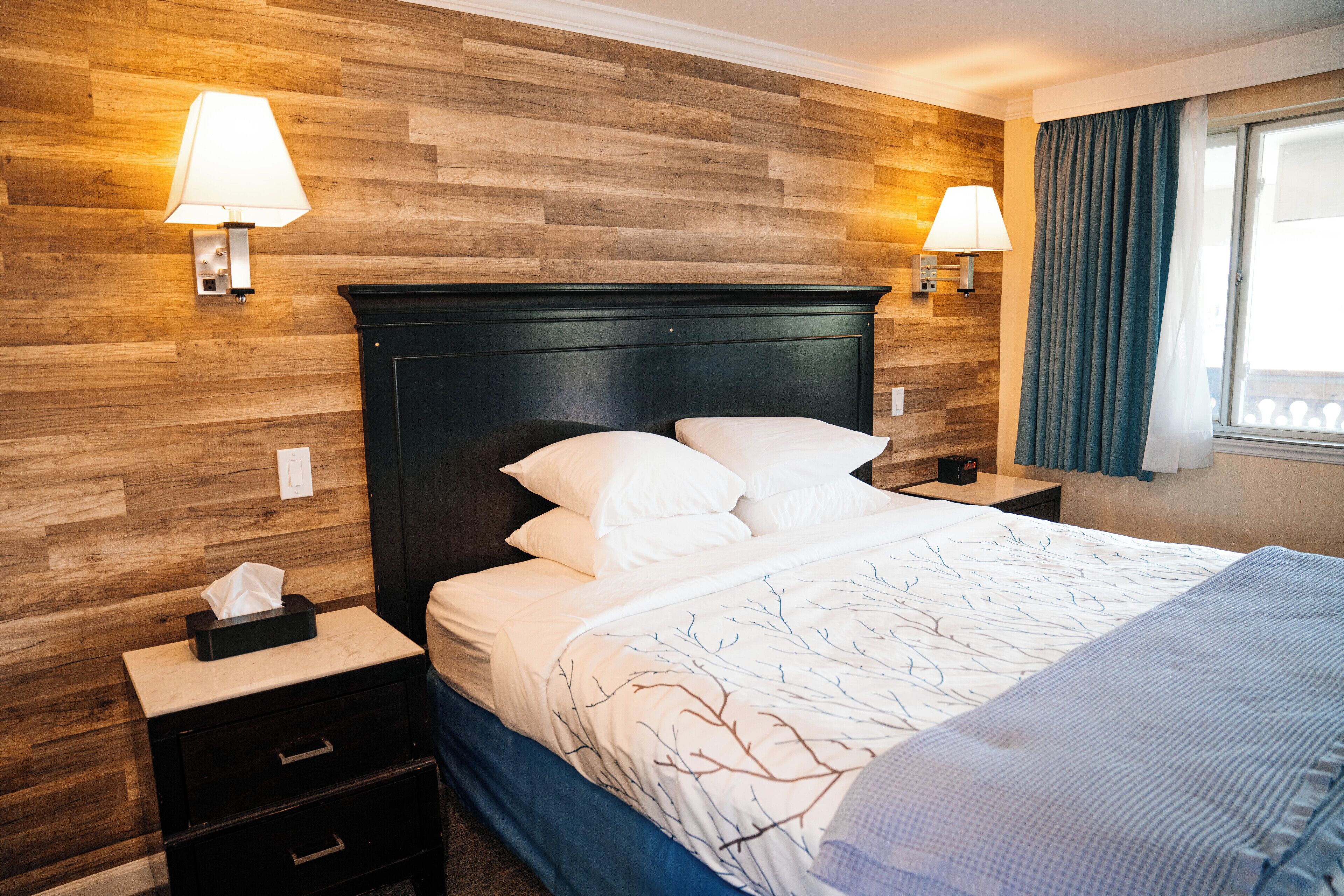 A neatly made bed with white linens and a blue blanket, a dark wooden headboard, two bedside lamps, and a nightstand with a tissue box.