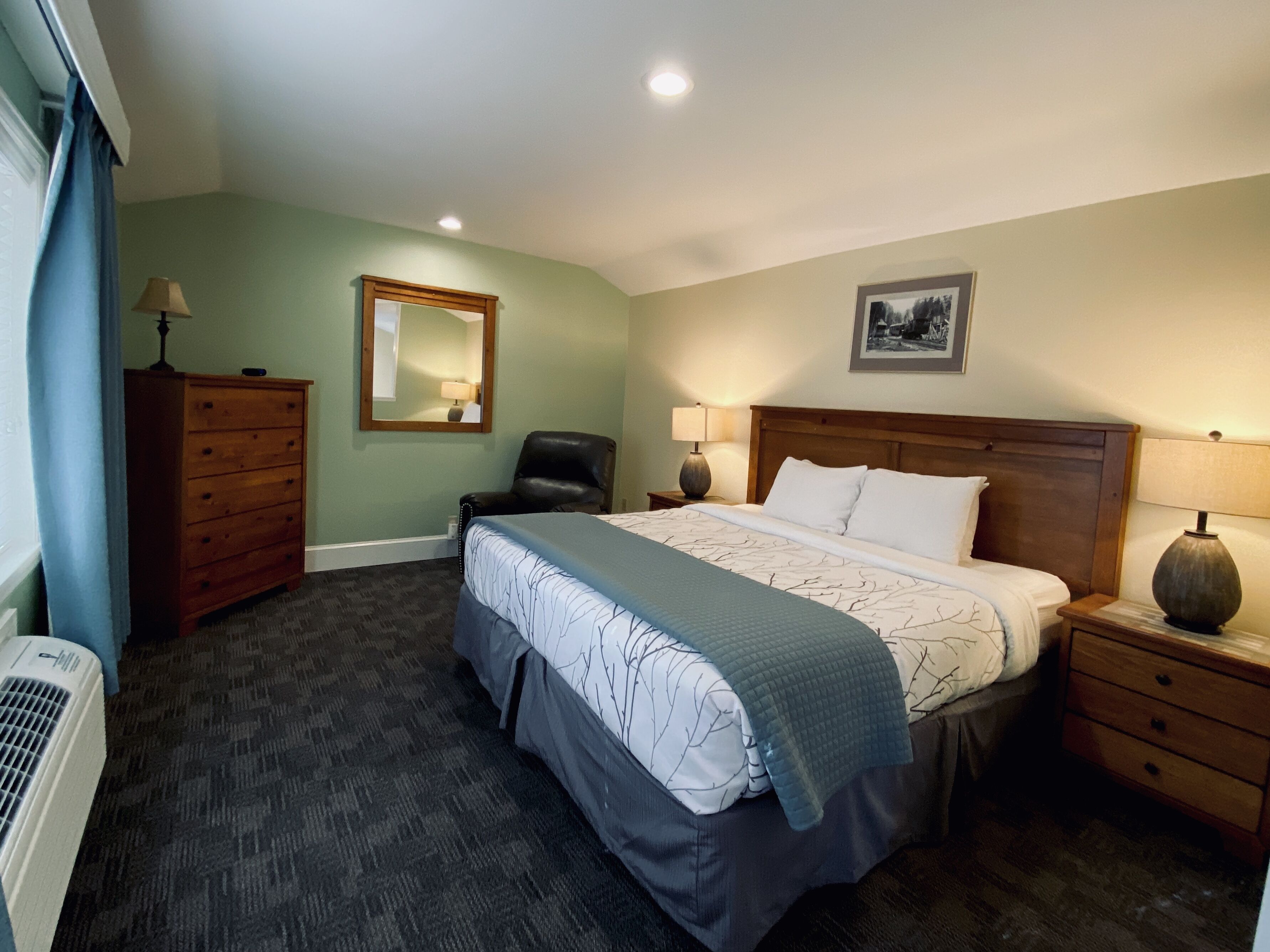Una habitación de hotel con una cama, una silla, un aparador, un espejo y un cuadro en la pared.