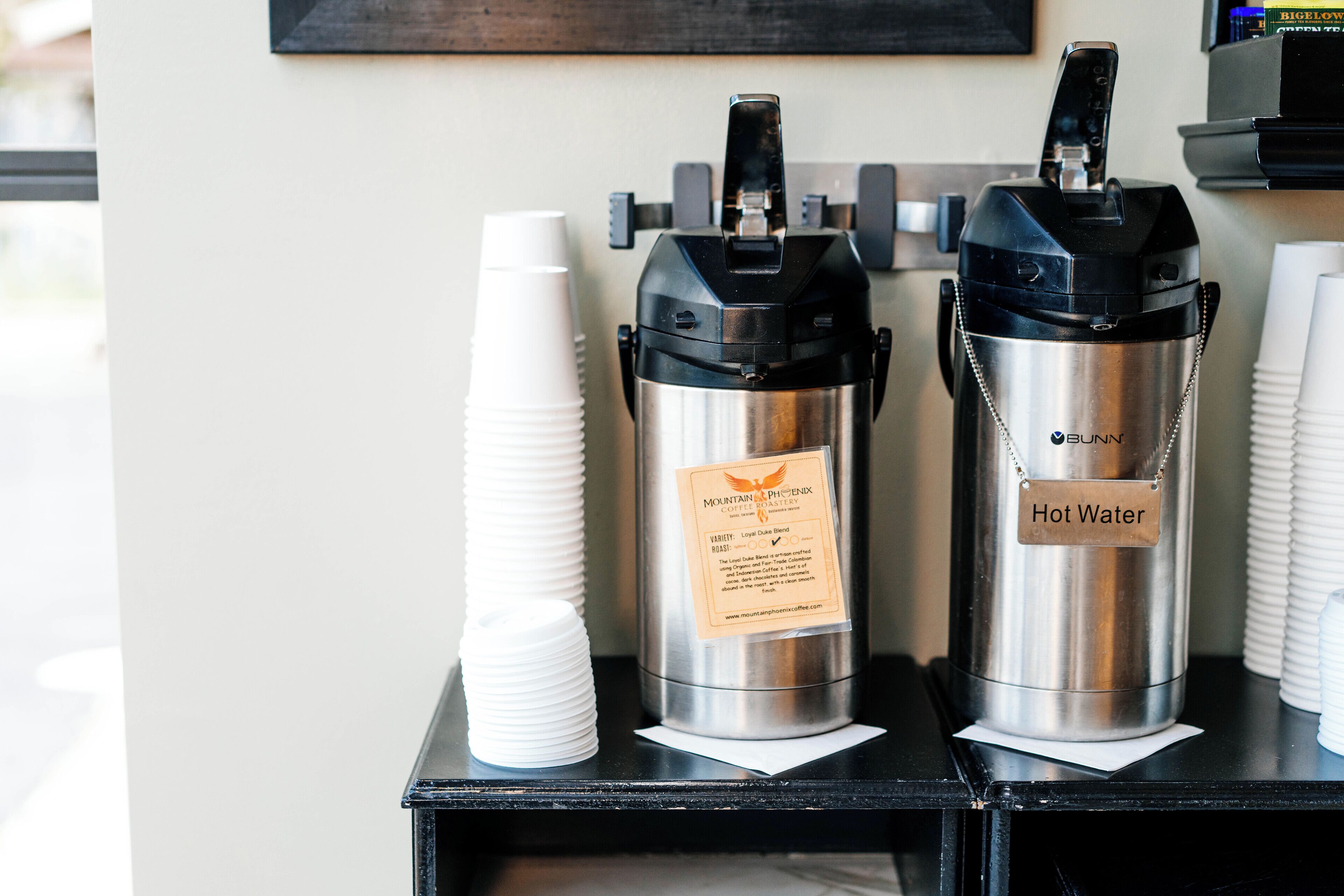 Two stainless steel hot water dispensers labeled "Bunn" and "Hot Water" on a black counter with white disposable cups stacked beside them.
