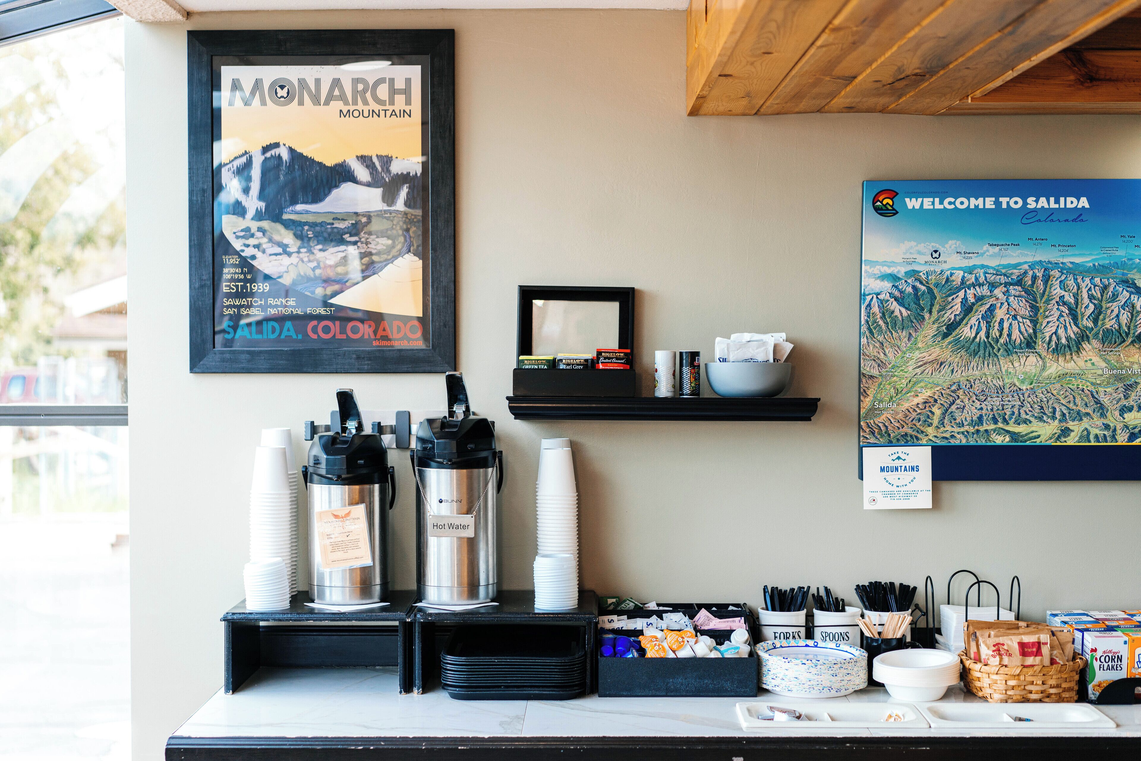 A hotel reception area with a framed poster of Monarch Mountain, a hot water dispenser, and a welcome sign for Salida.