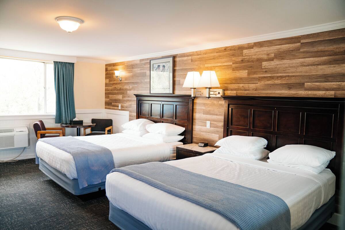 A hotel room with two beds, a desk, a chair, a window with curtains, and a wooden accent wall.