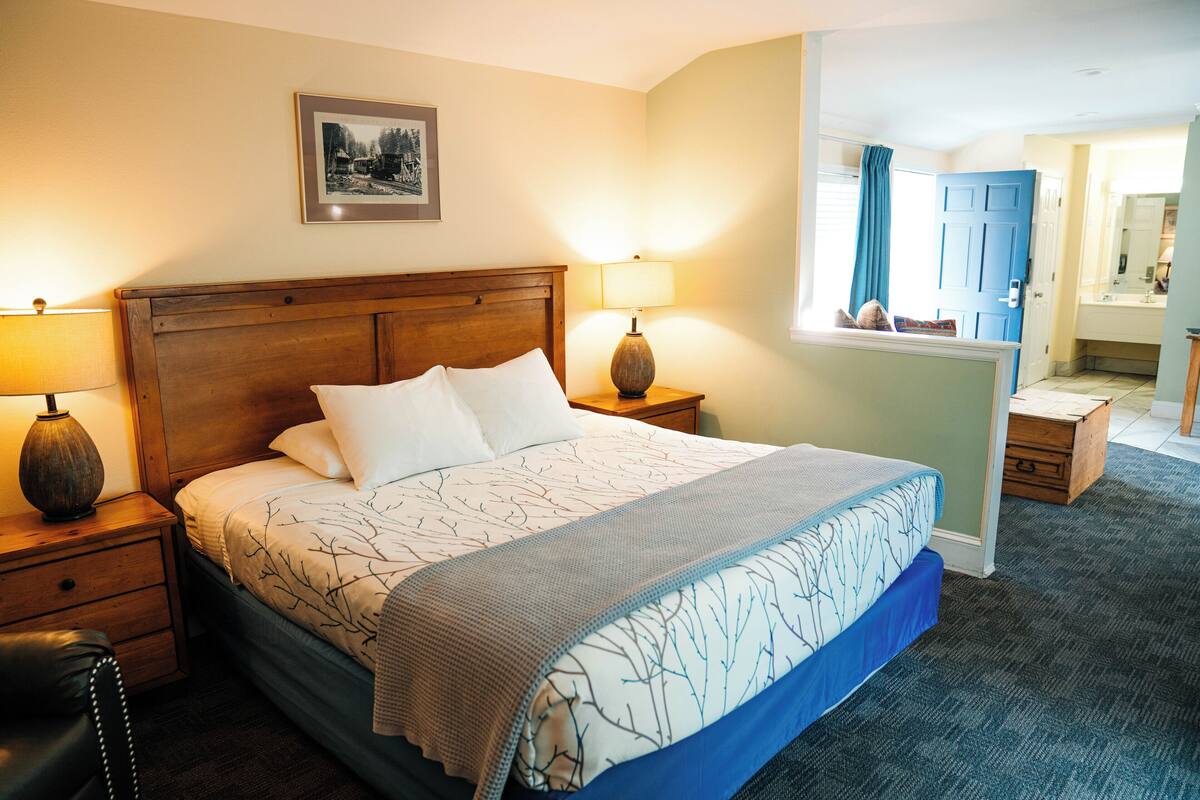 A neatly made bed with a patterned bedspread, two bedside lamps, a wooden nightstand, a small wooden chest, and a framed picture on the wall.