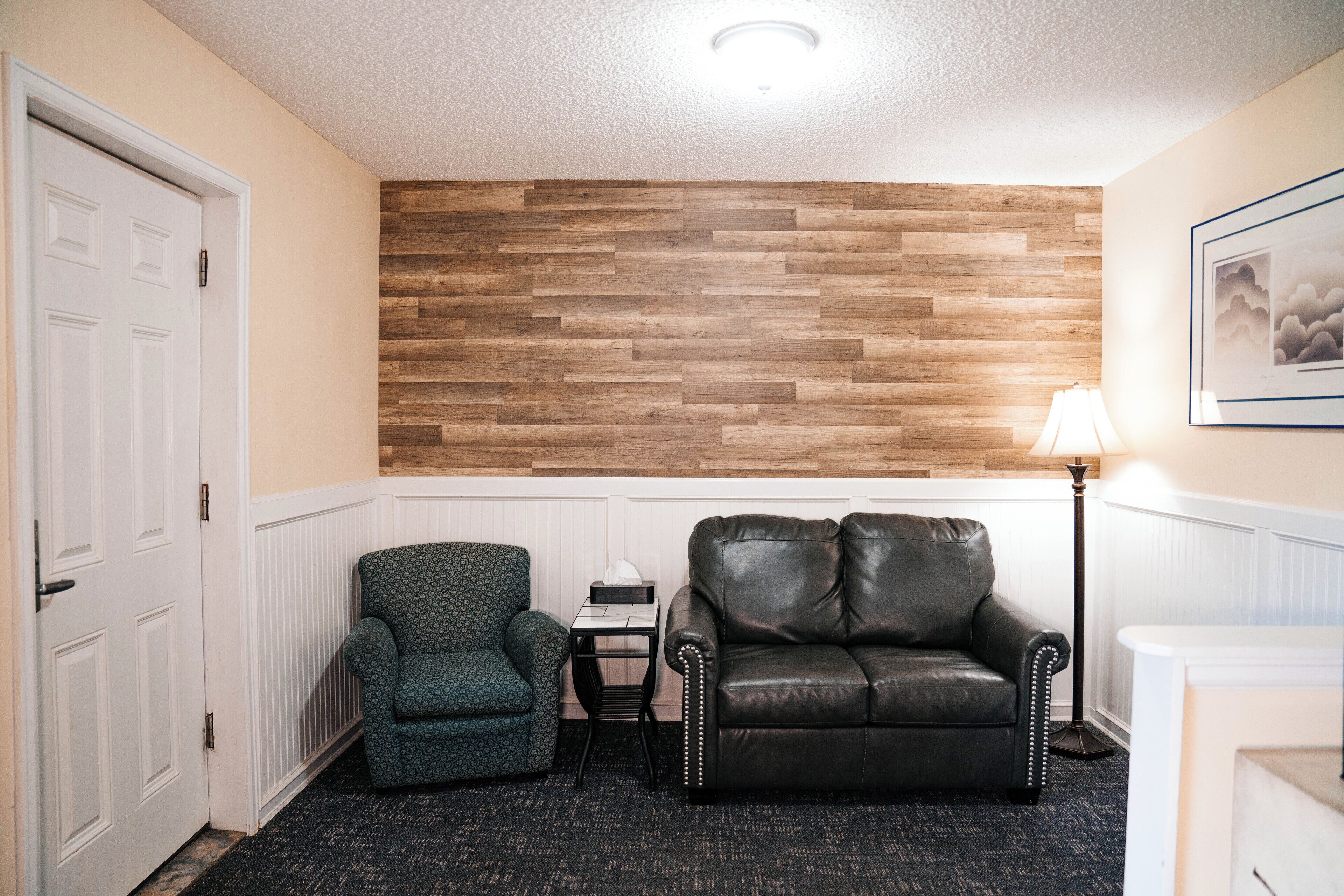 Una habitación con paneles de madera, un sofá de cuero negro, un sillón estampado, una mesita con una lámpara y una puerta blanca.