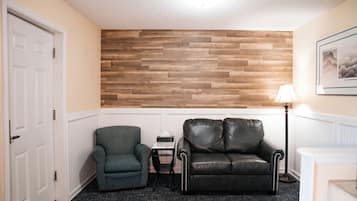 Una habitación con paneles de madera, un sofá de cuero negro, un sillón estampado, una mesita con una lámpara y una puerta blanca.