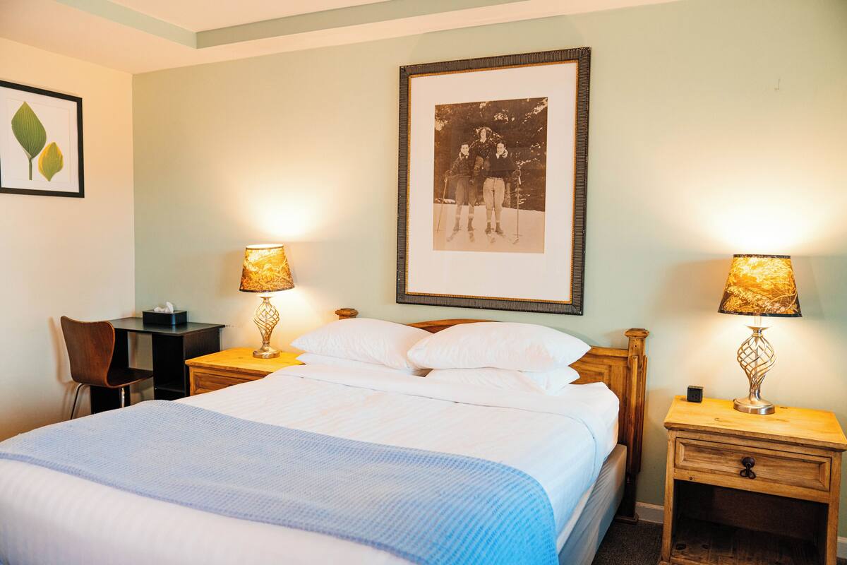 A neatly made bed with white linens and a blue blanket, flanked by two bedside tables with lamps, a framed photograph on the wall, and a chair to the left.