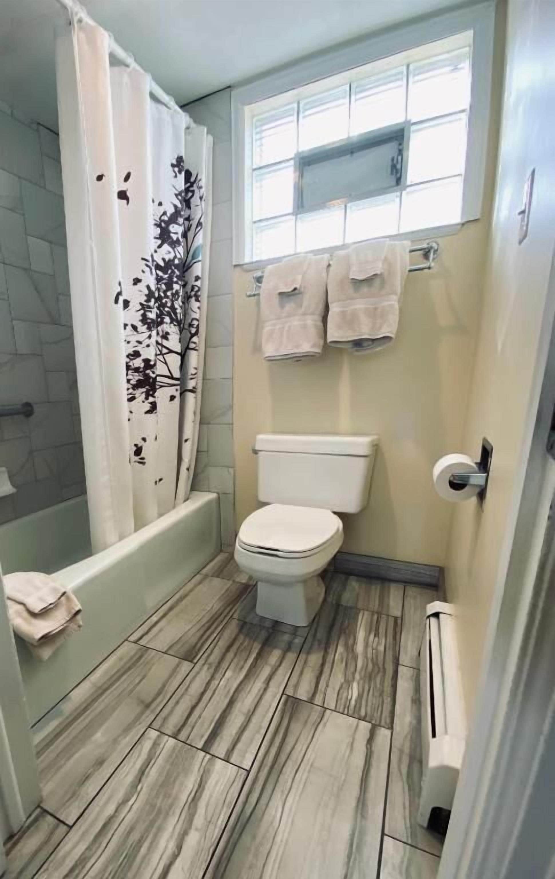 A bathroom with a toilet, a shower with a tree-patterned curtain, and a window above the toilet.