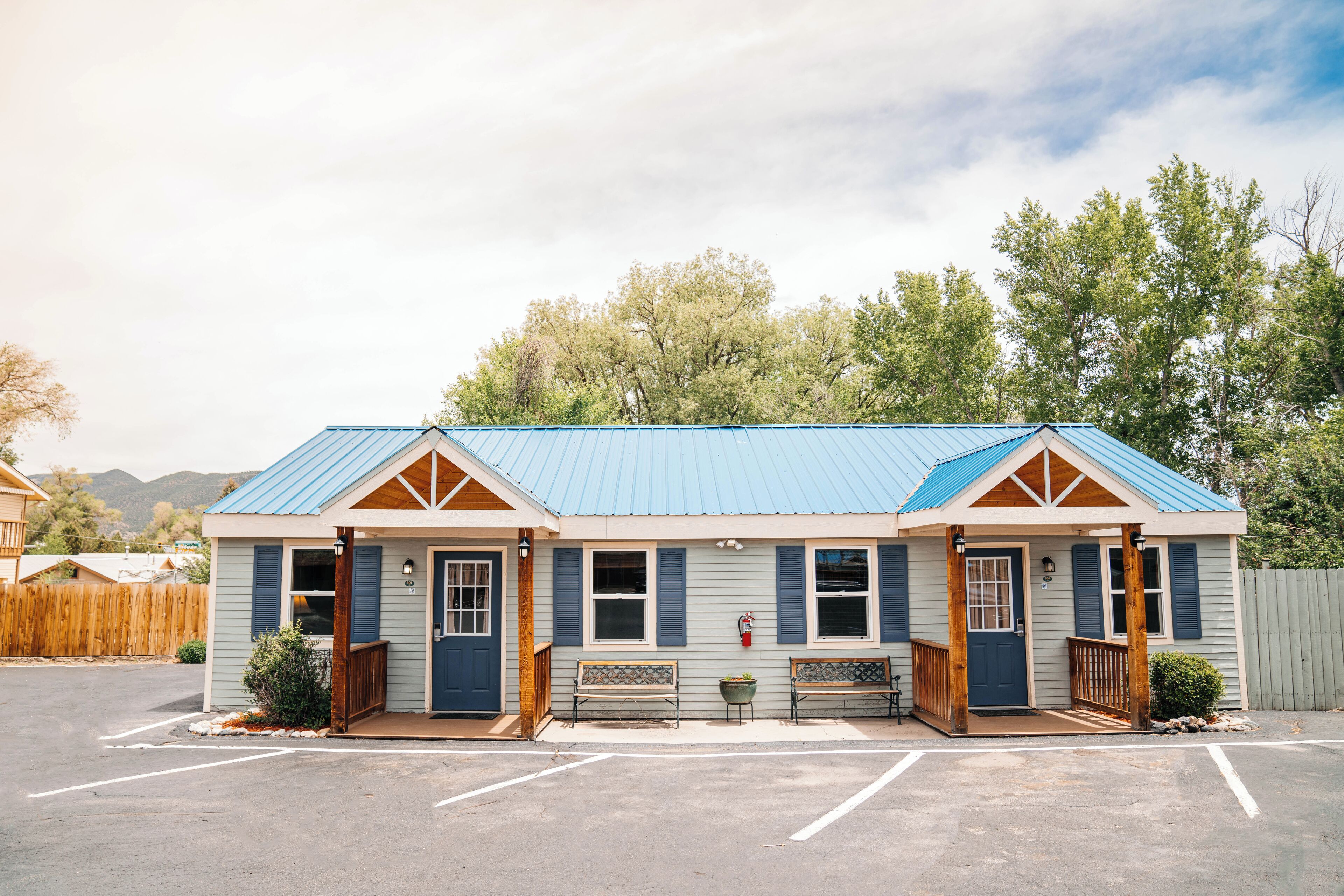 A small, single-story building with a blue roof and wooden trim, featuring two doors, a bench, and a small porch area.