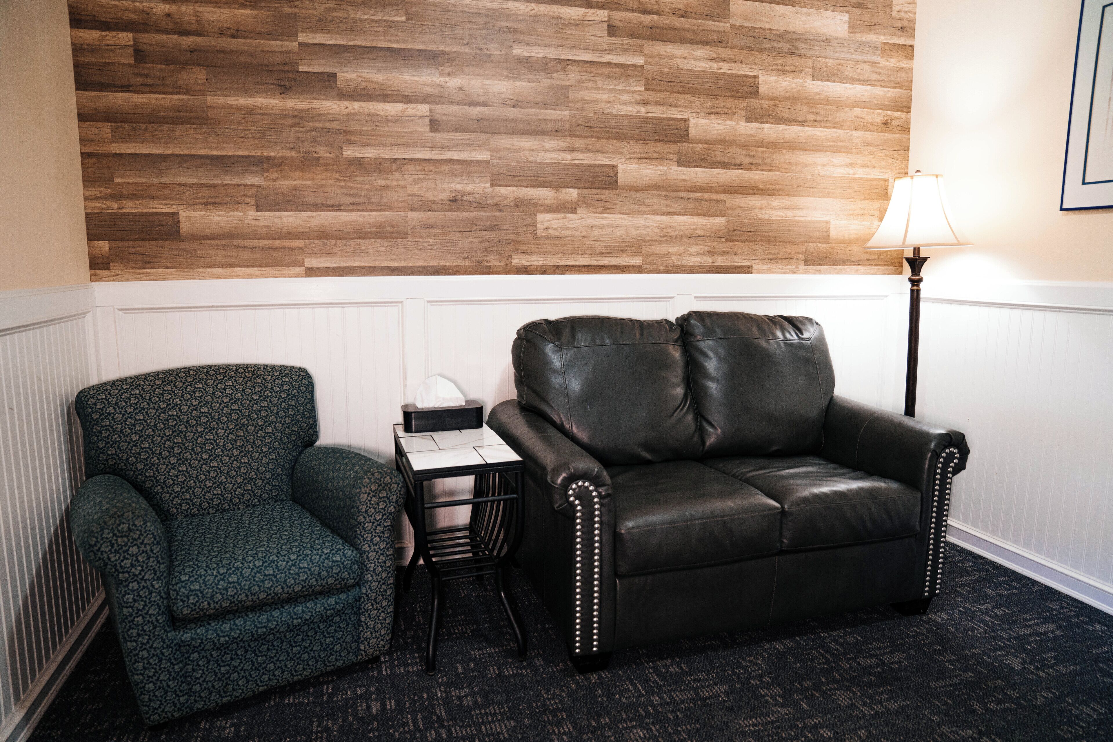 A room with a wooden wall, a black leather sofa, a patterned armchair, a small table with a tissue box, and a floor lamp.