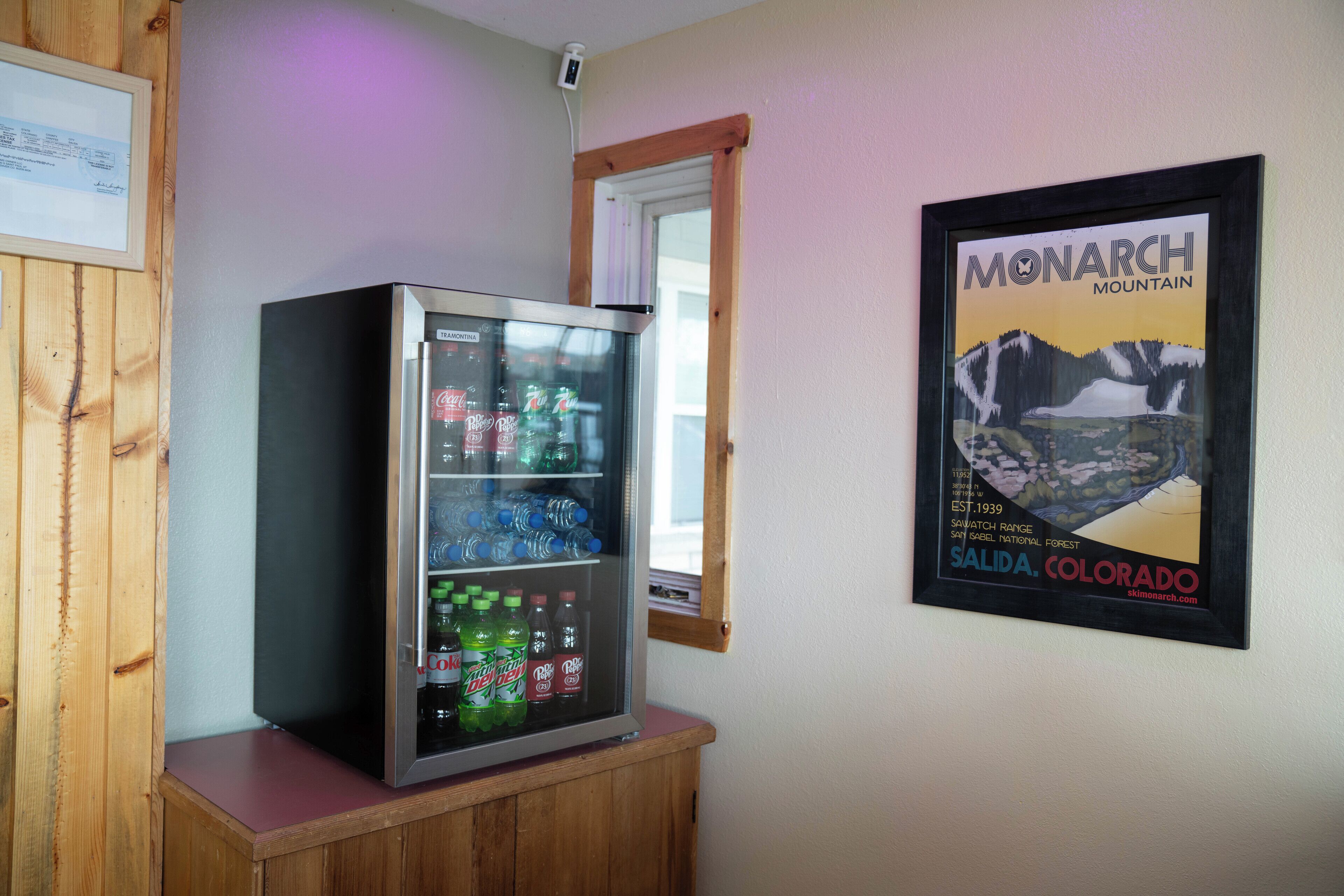 A refrigerator stocked with beverages, a framed poster of Monarch Mountain, and a wooden cabinet.