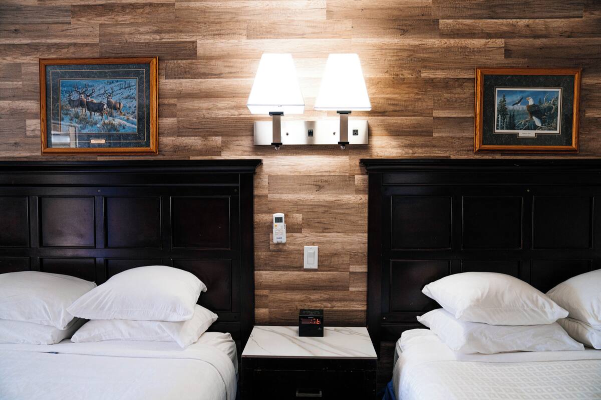 A hotel room with two beds, a nightstand with a clock, wall-mounted lamps, and framed pictures on the wooden wall.