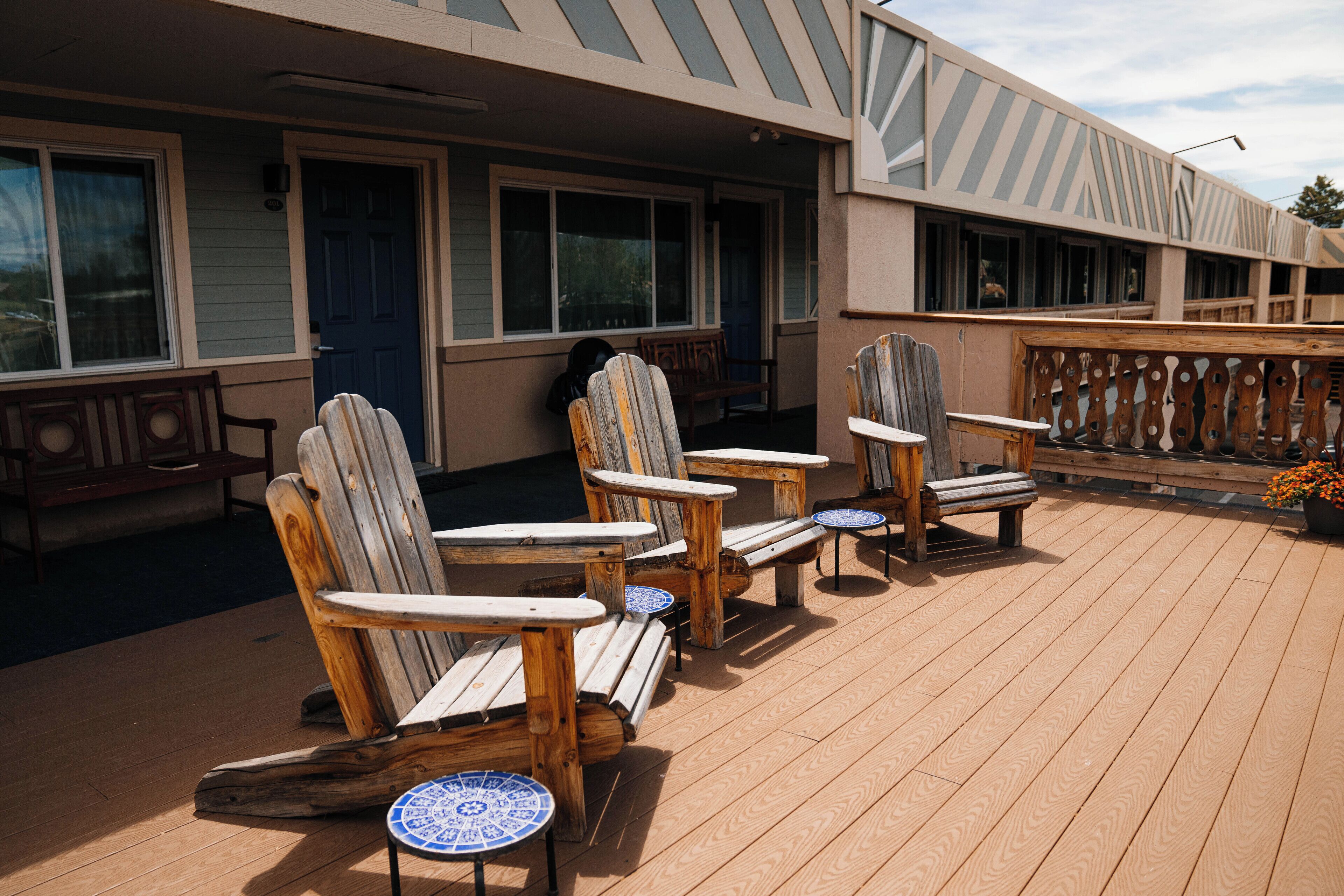 A wooden deck with Adirondack chairs and small tables, a covered porch, and a building with large windows and a blue door.