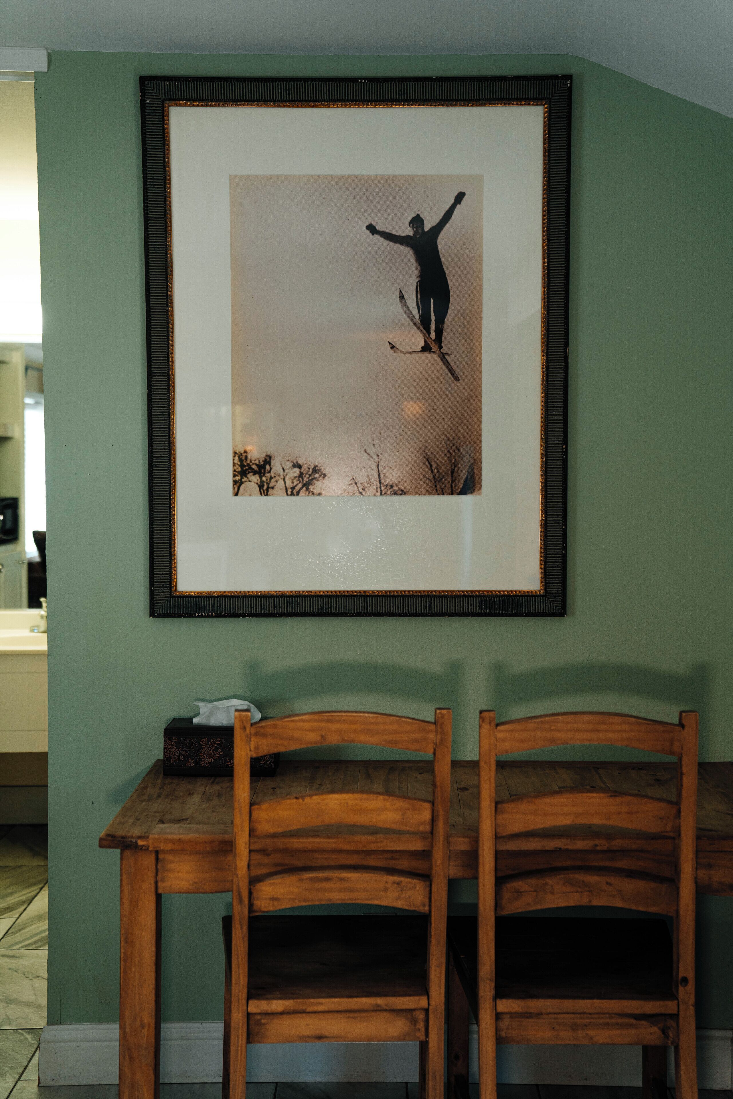 A framed picture of a person skiing on a mountain, a wooden table, and two wooden chairs.
