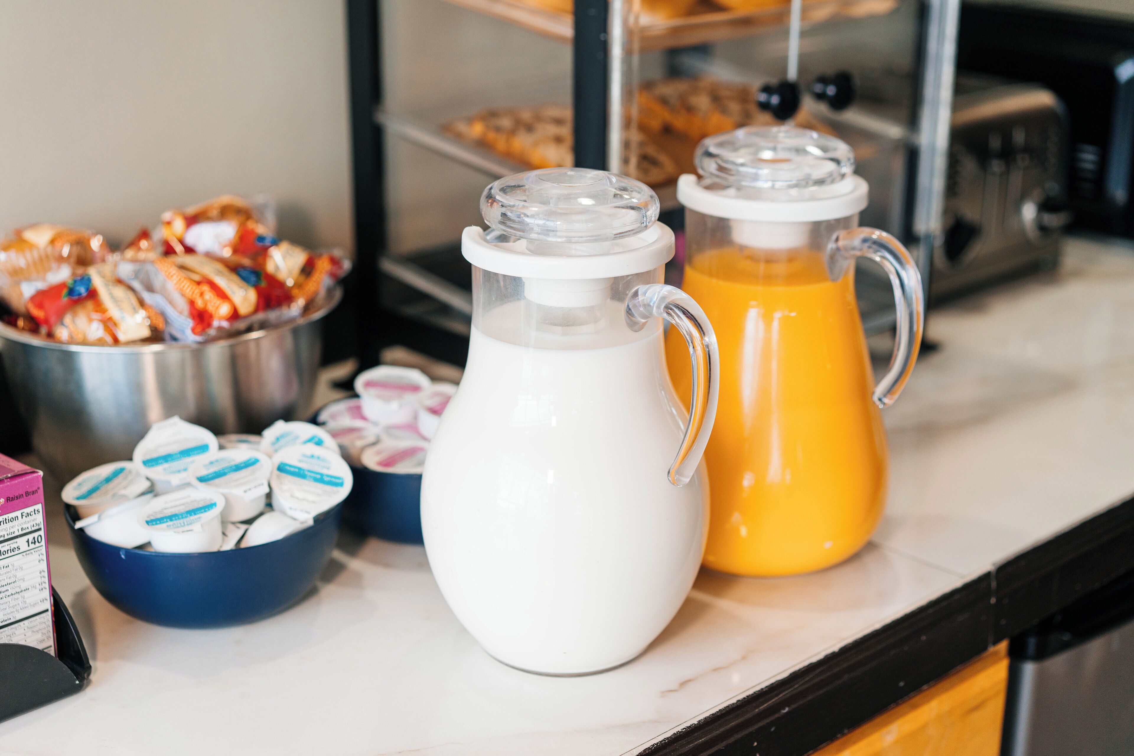 A breakfast buffet with yogurt cups, a milk pitcher, and a juice pitcher on a marble countertop.