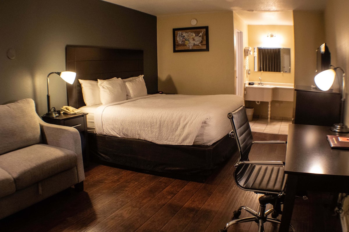 Ein Hotelzimmer mit Bett, Schreibtisch, Sessel, Sofa, Nachttisch mit Lampe, Telefon, Badezimmer mit Waschbecken und Spiegel sowie einer Wanduhr.