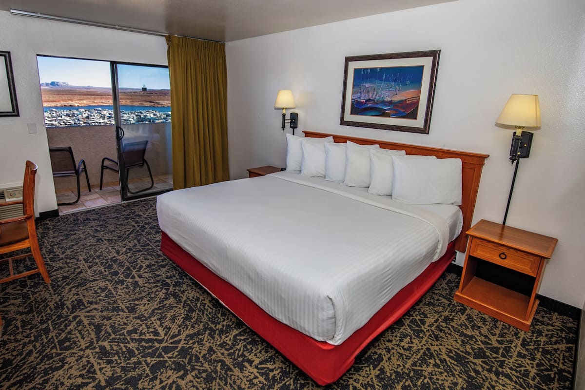 A hotel room with a large bed, two bedside lamps, a wooden nightstand, a chair, a large window with a view of a cityscape, and a framed picture on the wall.