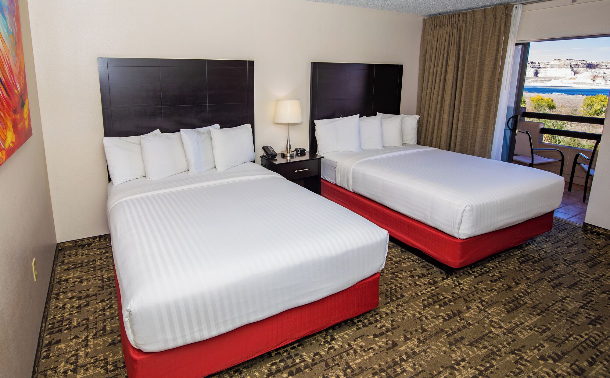 A hotel room with two beds, a nightstand, a lamp, and a view of the outdoors.