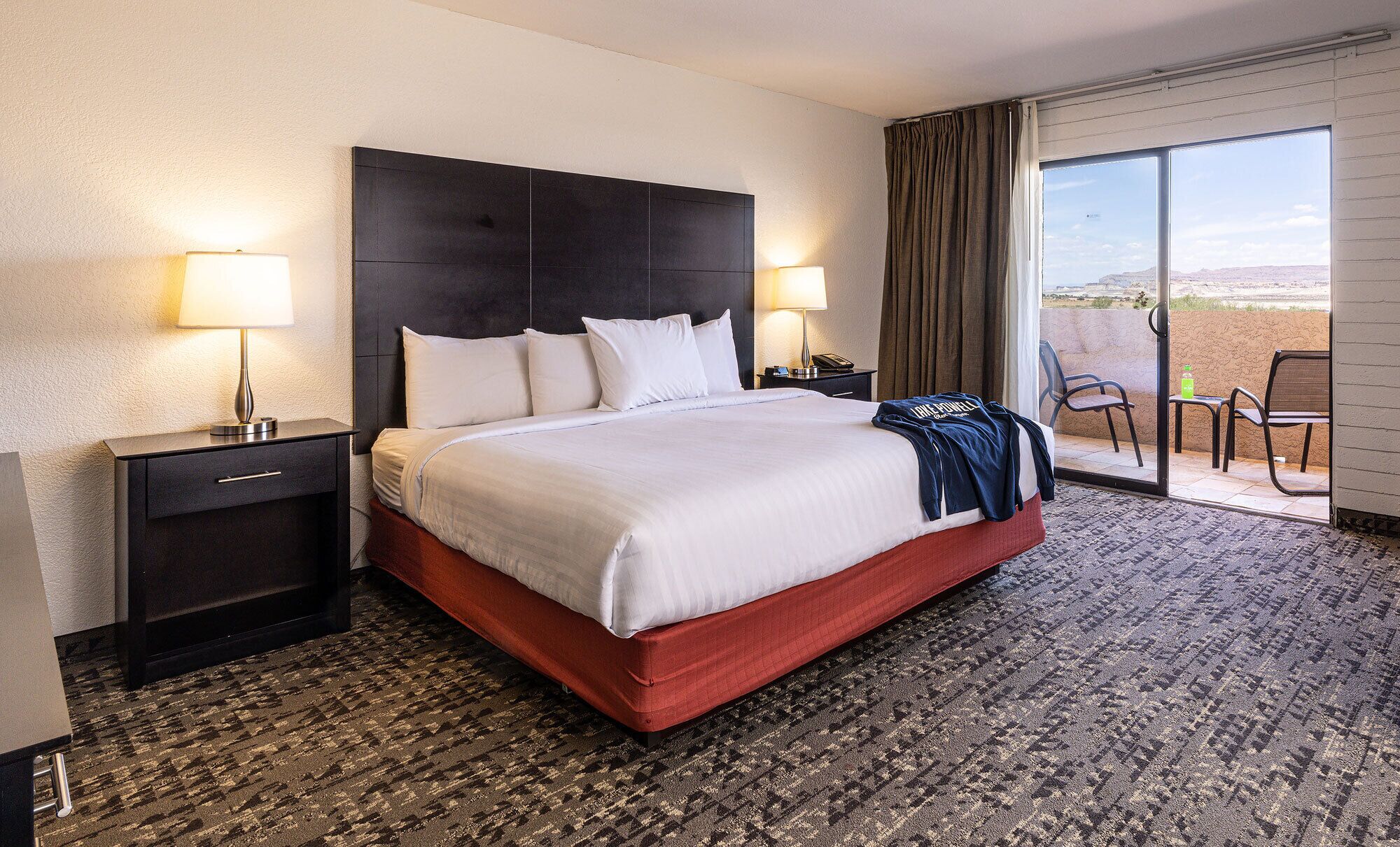 A hotel room with a large bed, two bedside tables with lamps, a red upholstered headboard, a carpeted floor, and a view of the desert through a sliding glass door.