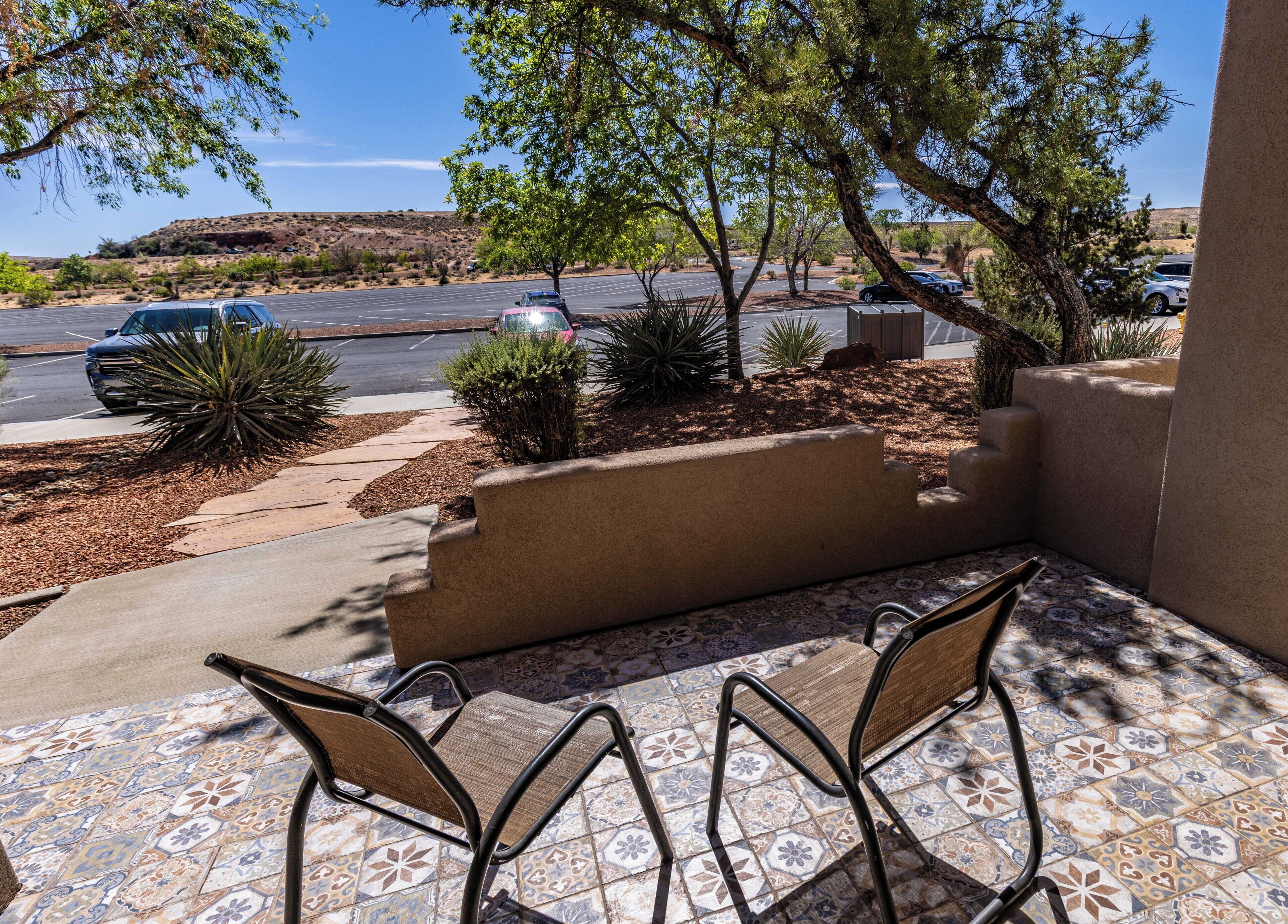 A patio with two chairs, a tiled floor, and a view of a parking lot with cars and a hilly landscape.
