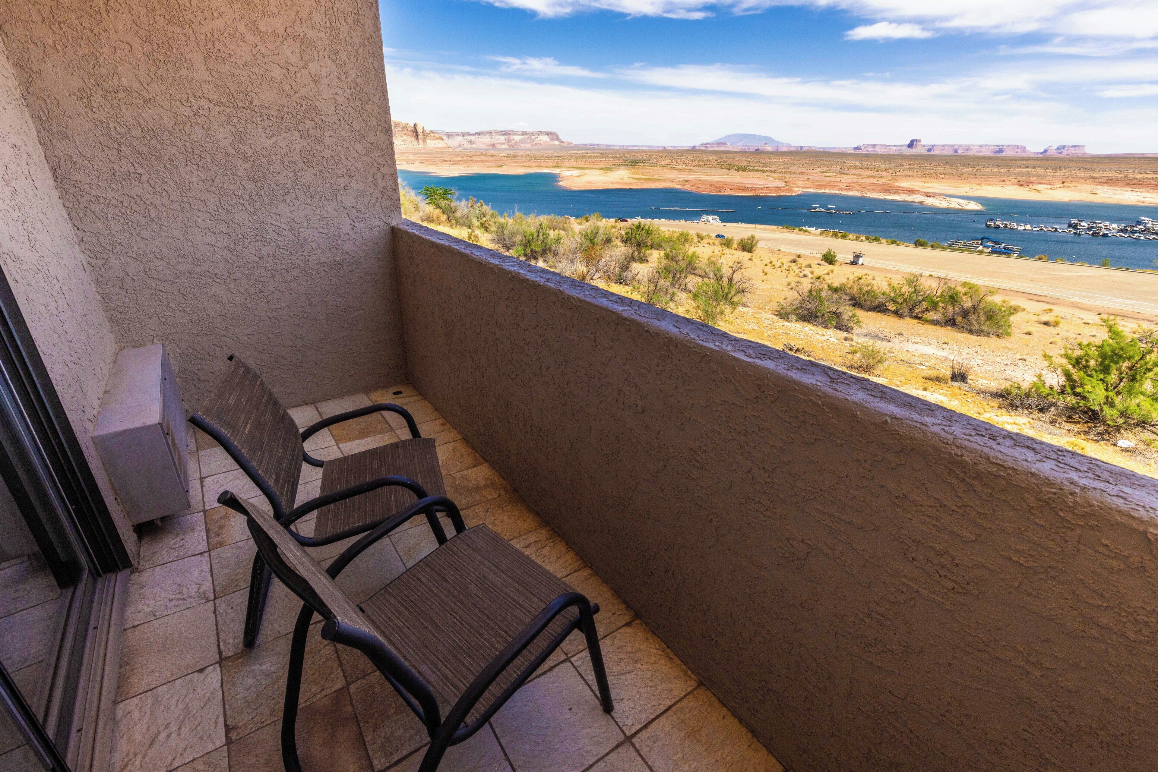 Ein Balkon mit zwei Stühlen und einem Tisch, der auf eine Wüstenlandschaft mit einem Gewässer und fernen Bergen blickt.