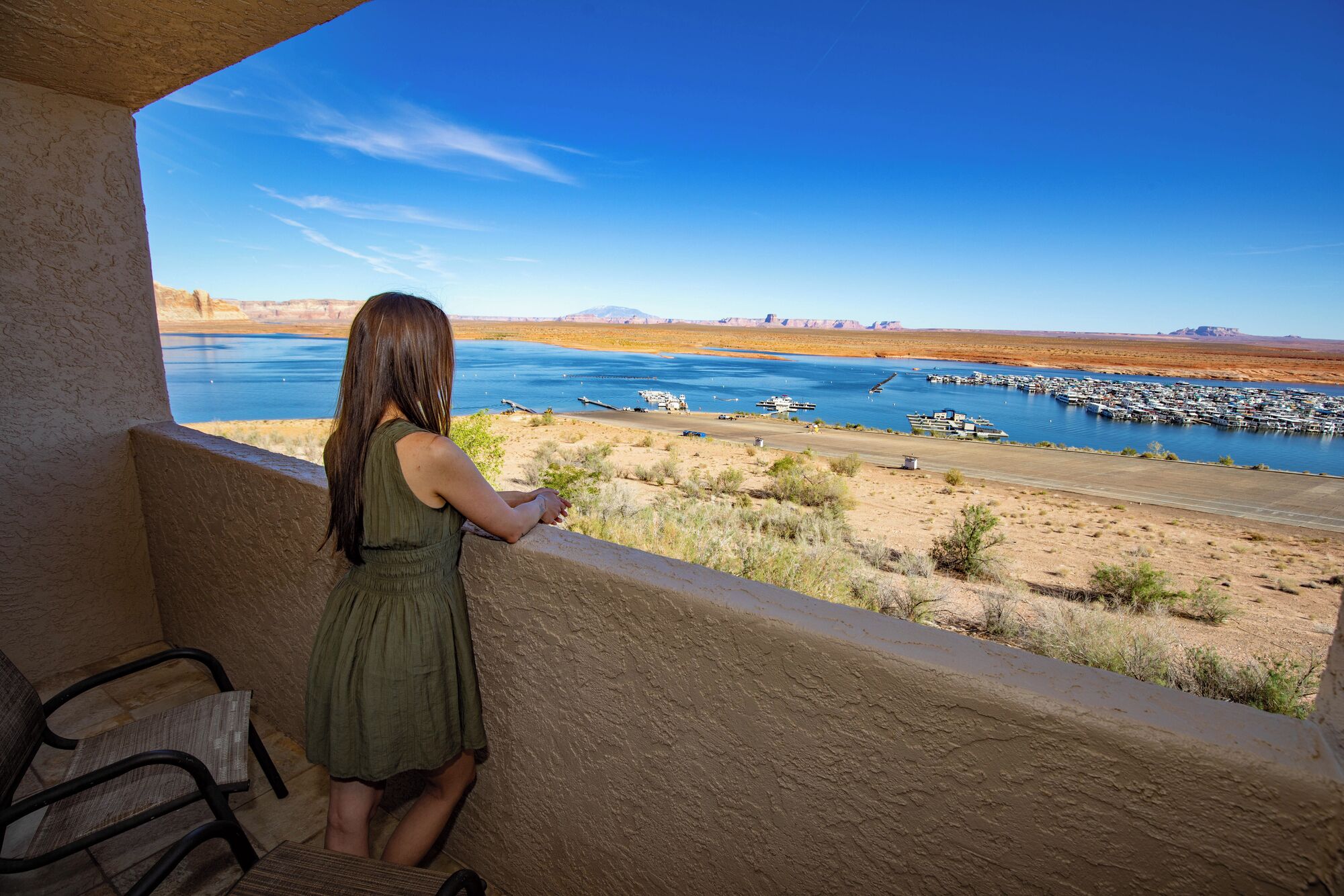 A woman in a green dress standing by a window overlooking a marina with boats and a desert landscape.