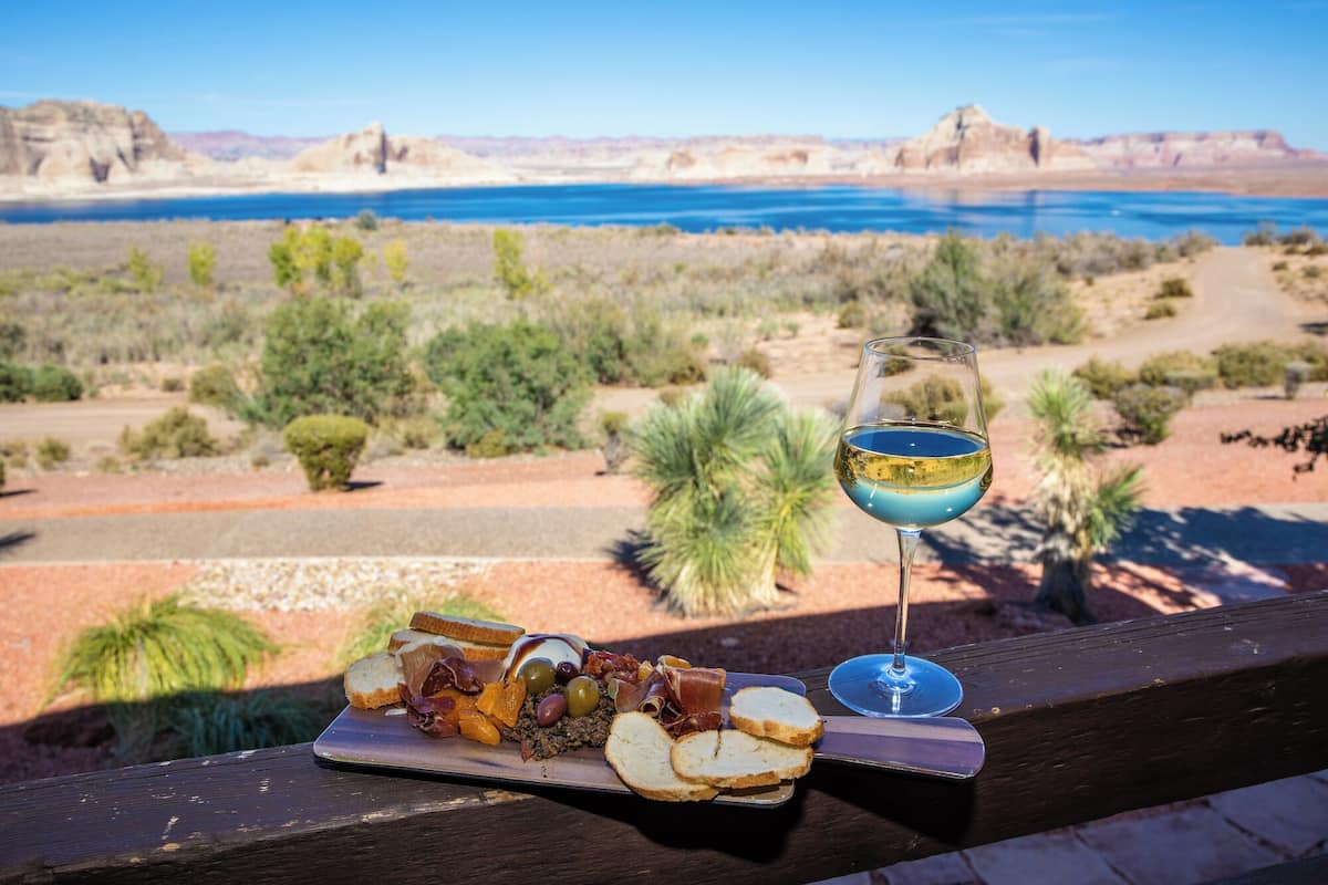 A meal with bread, cheese, olives, and meats on a tray, with a glass of blue drink beside it, overlooking a desert landscape with a body of water and mountains in the background.