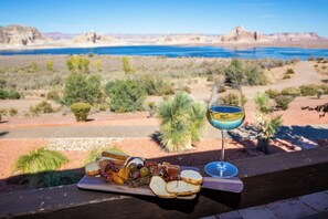 Una bandeja con comida que incluye pan, queso, aceitunas y embutidos, junto a un vaso de bebida azul, con vistas a un paisaje desértico que incluye un cuerpo de agua y montañas al fondo.