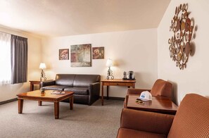 A hotel room with a leather sofa, a wooden coffee table, a wicker armchair, and a desk with a lamp and a coffee maker.