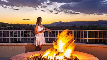 Una mujer de pie junto a un hogar de piedra con un sombrero encima, contemplando un paisaje desértico al atardecer.