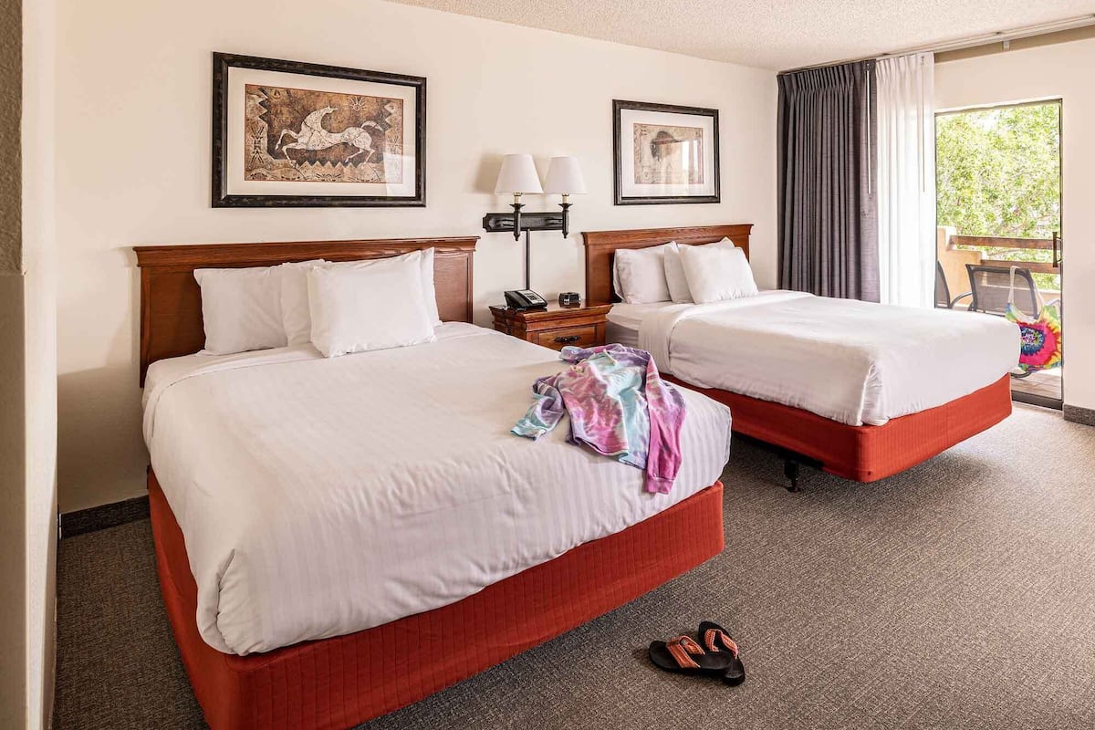 A hotel room with two beds, a nightstand, a lamp, and a window with curtains.