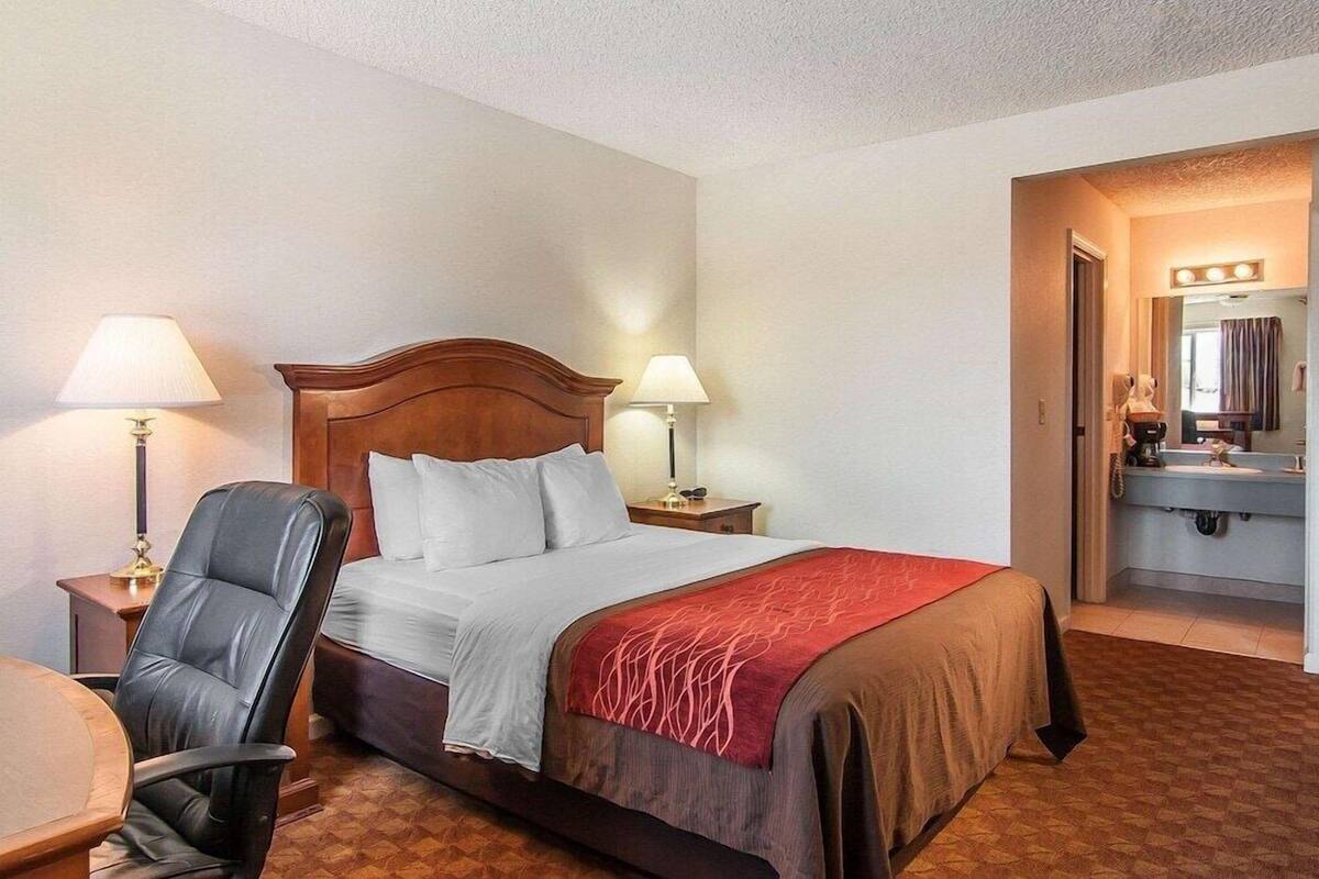 A hotel room with a bed, two bedside lamps, a chair, a desk, and a bathroom with a mirror and sink.