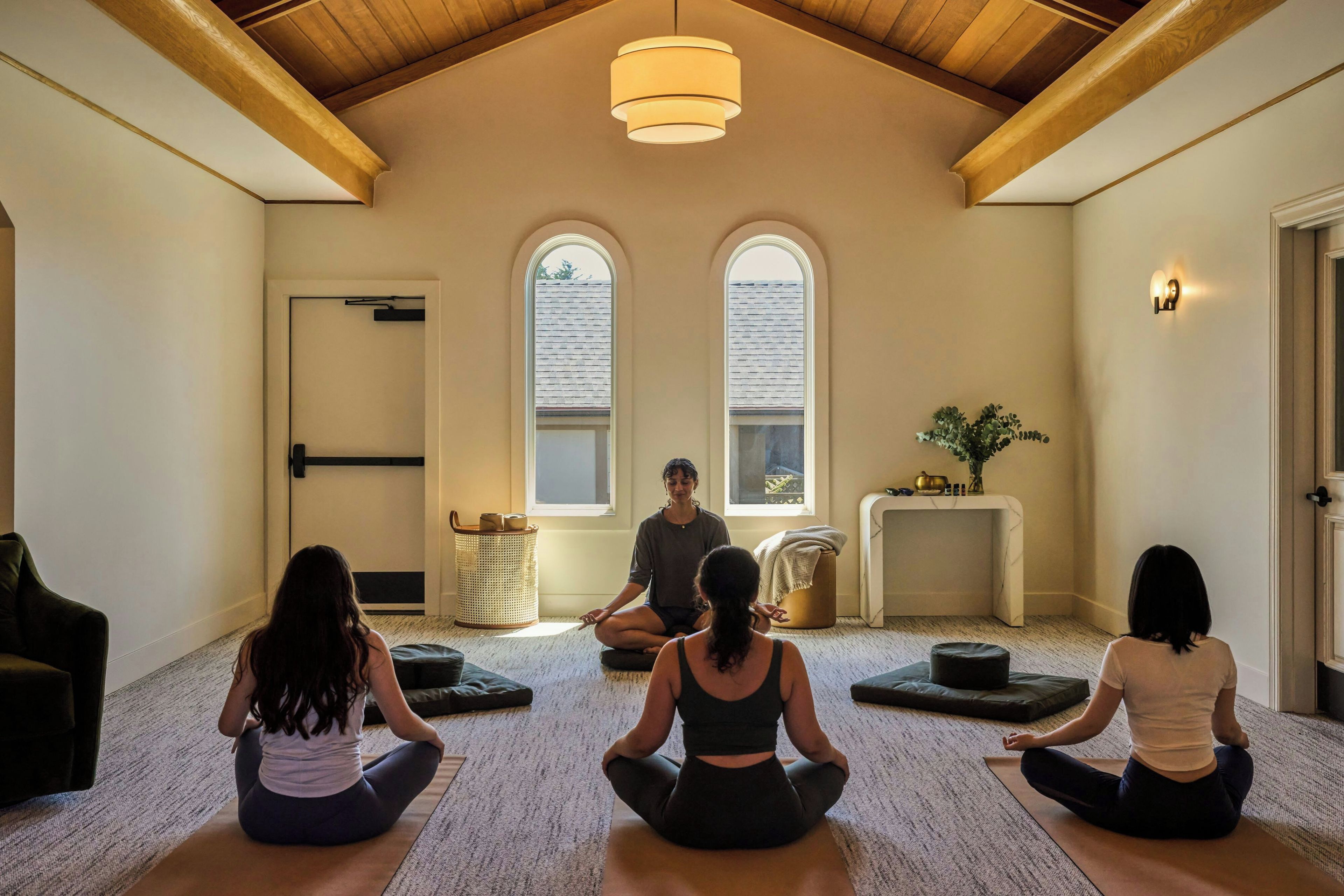 A yoga class in a room with a high ceiling, two windows, and a ceiling light.