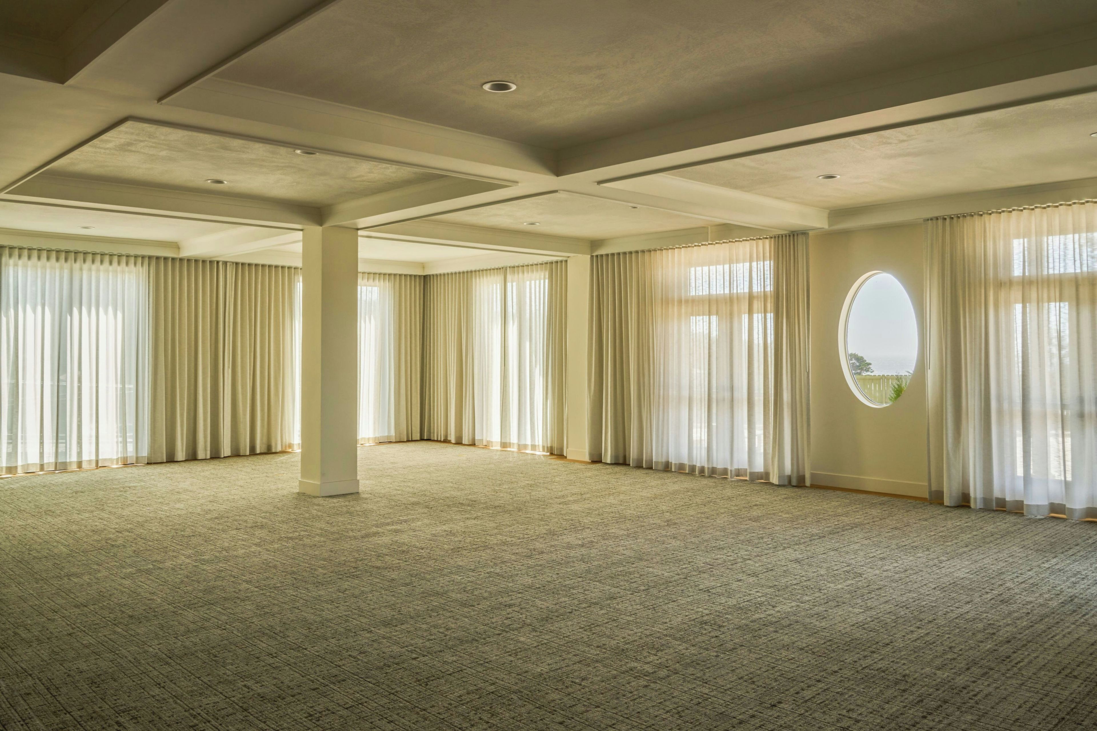 A spacious room with a high ceiling, large windows with sheer curtains, and a carpeted floor.
