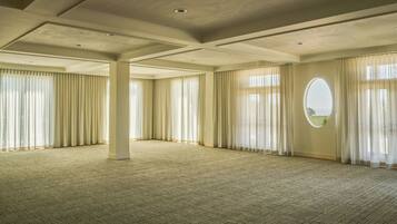 A spacious room with a high ceiling, large windows with sheer curtains, and a carpeted floor.