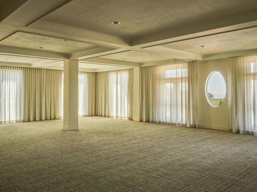 A spacious room with a high ceiling, large windows with sheer curtains, and a carpeted floor.