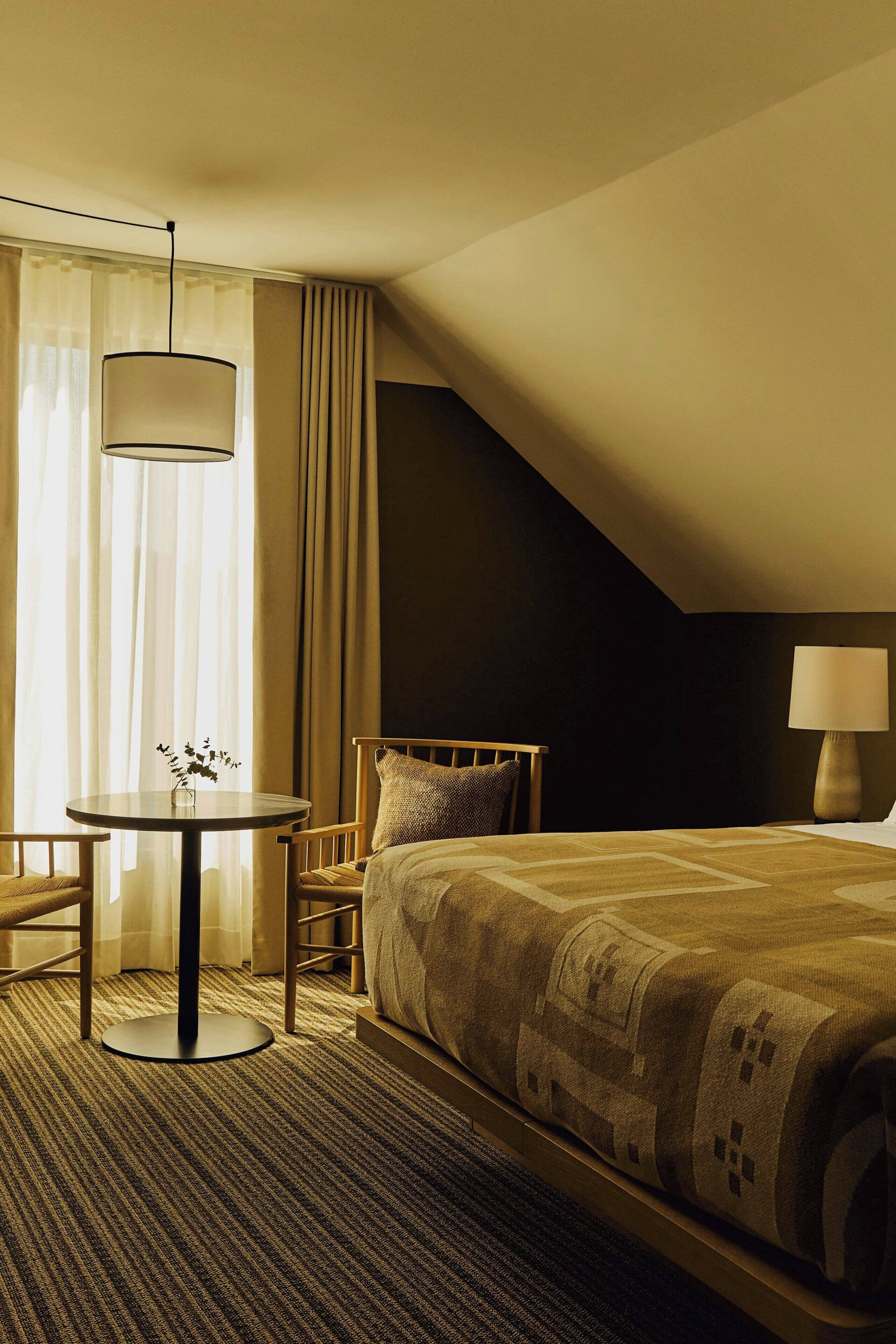 Habitación de hotel con cama, una mesa redonda con un florero, una silla, una lámpara y una ventana con cortinas.