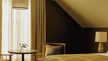 Habitación de hotel con cama, una mesa redonda con un florero, una silla, una lámpara y una ventana con cortinas.