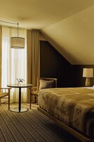 Habitación de hotel con cama, una mesa redonda con un florero, una silla, una lámpara y una ventana con cortinas.