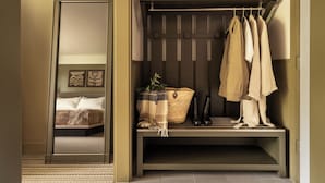 A room with a bed, a bench with hanging clothes, a basket, and a shelf with books.