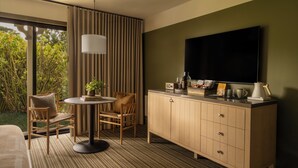 A hotel room with a large window, a round table, two chairs, a cabinet with a flat-screen TV, and a coffee maker.