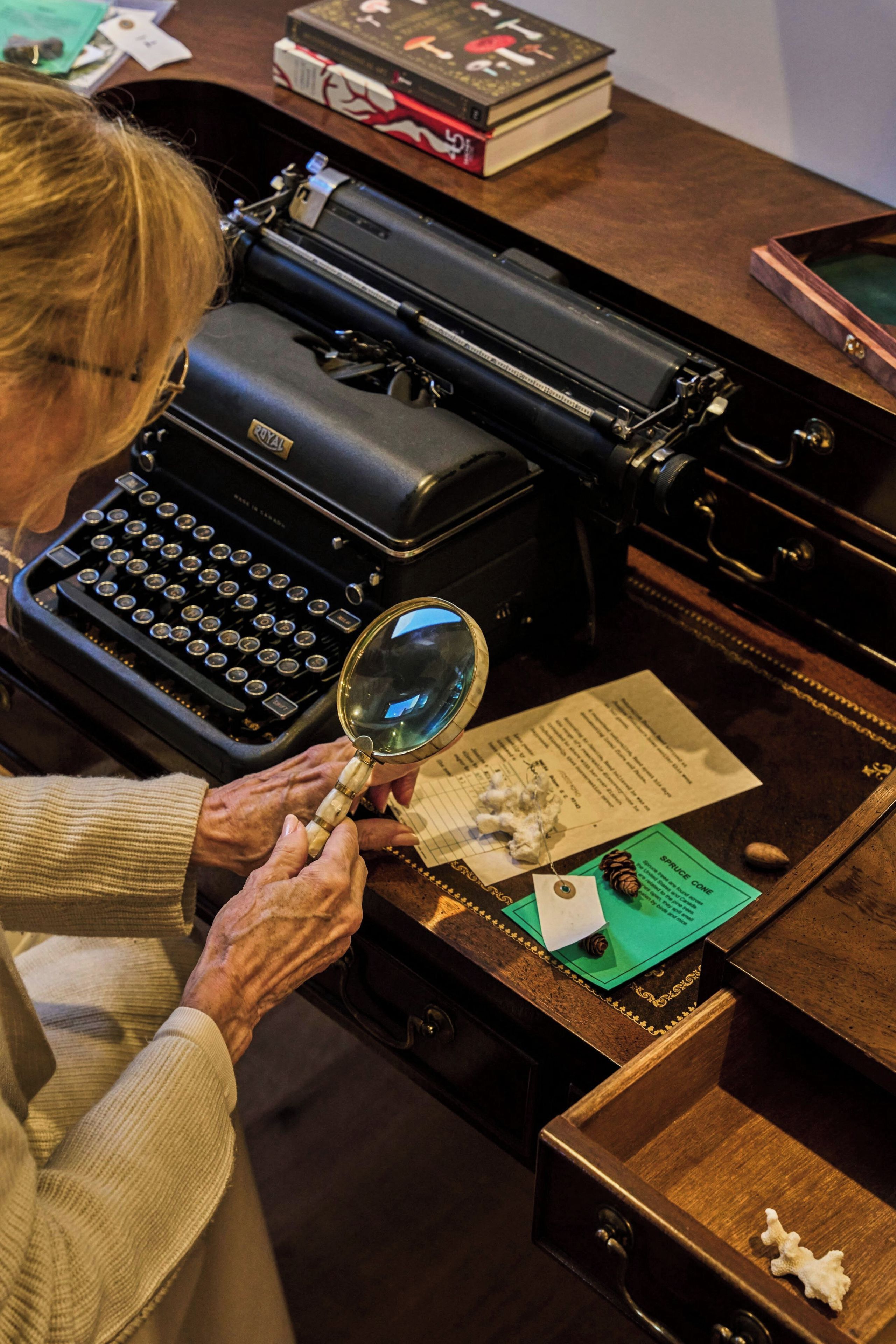 A person using a magnifying glass over a document on a desk with a typewriter and a book.