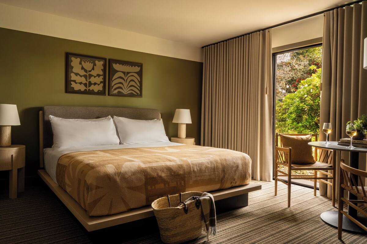 Une chambre d’hôtel avec un lit, deux lampes de chevet, une petite table avec une chaise, et une vue sur la verdure par la fenêtre.