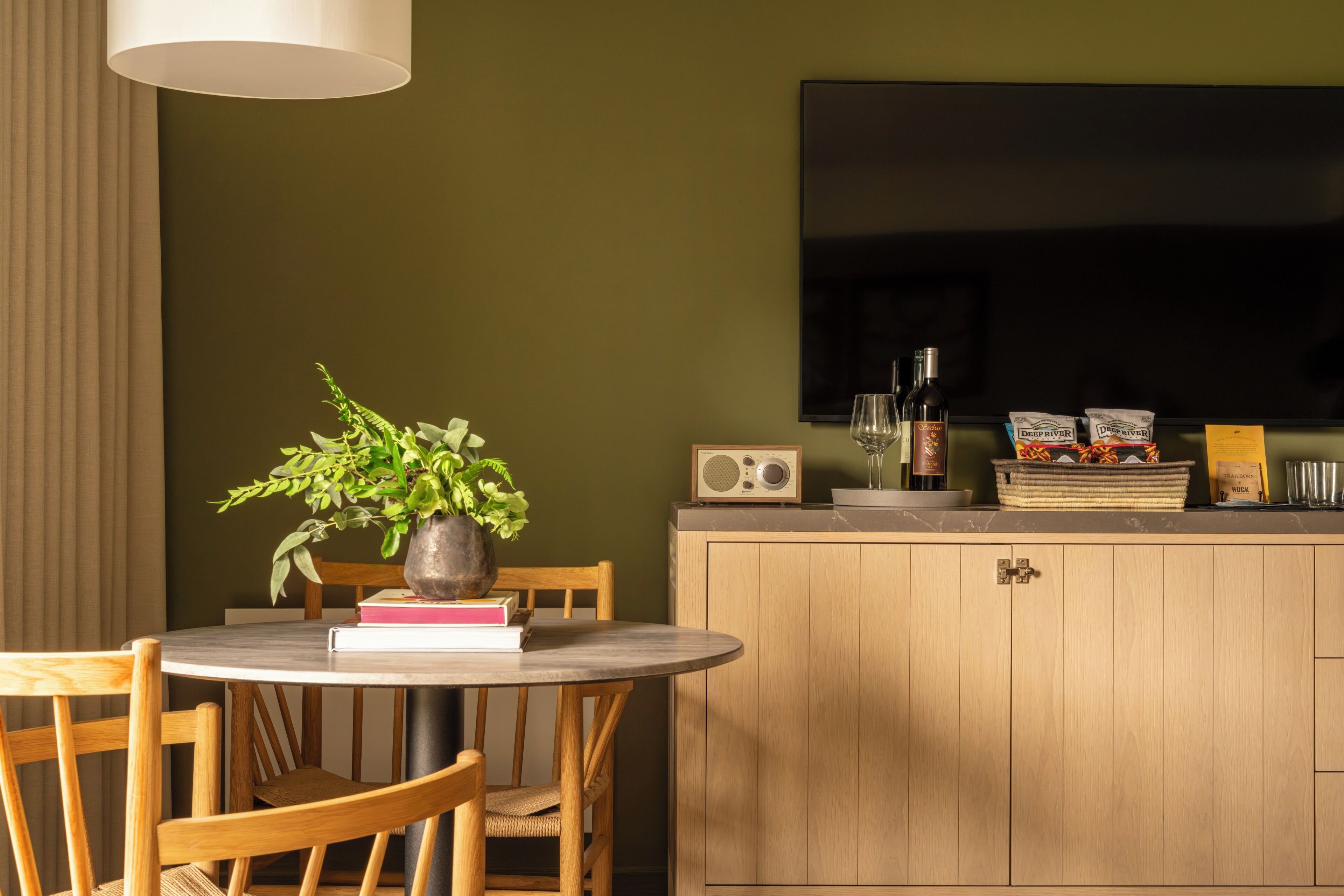 Un comedor con una mesa redonda, sillas, una planta en maceta, libros, un televisor y una encimera de cocina con objetos.