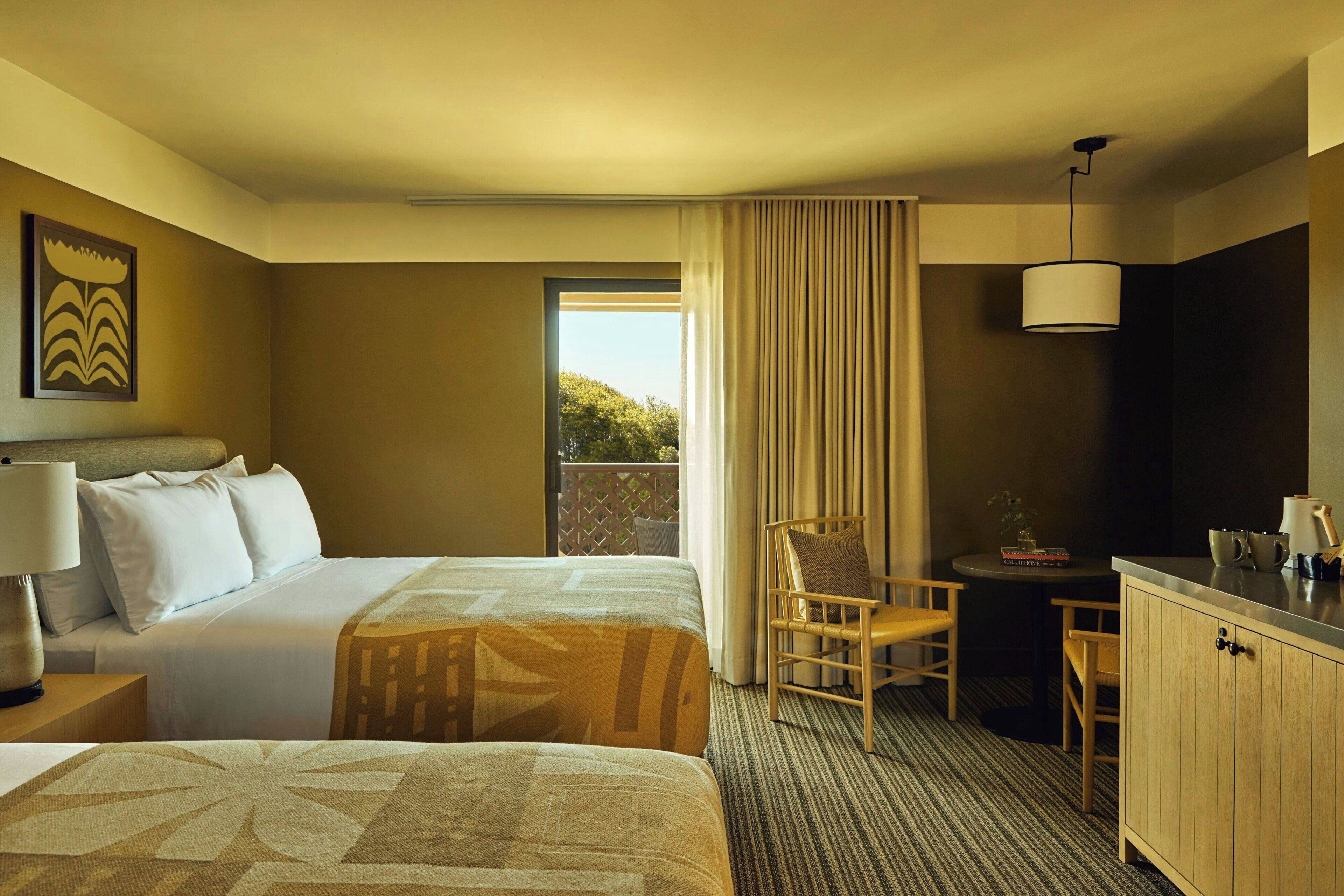 Habitación de hotel con dos camas, un escritorio, una silla, una lámpara y vistas al exterior.