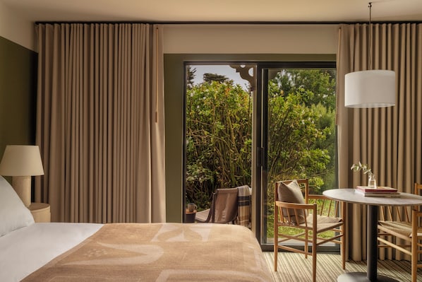 Une chambre d’hôtel avec un lit, une table ronde entourée de chaises, une lampe et une vue sur la verdure, visible à travers une porte vitrée.