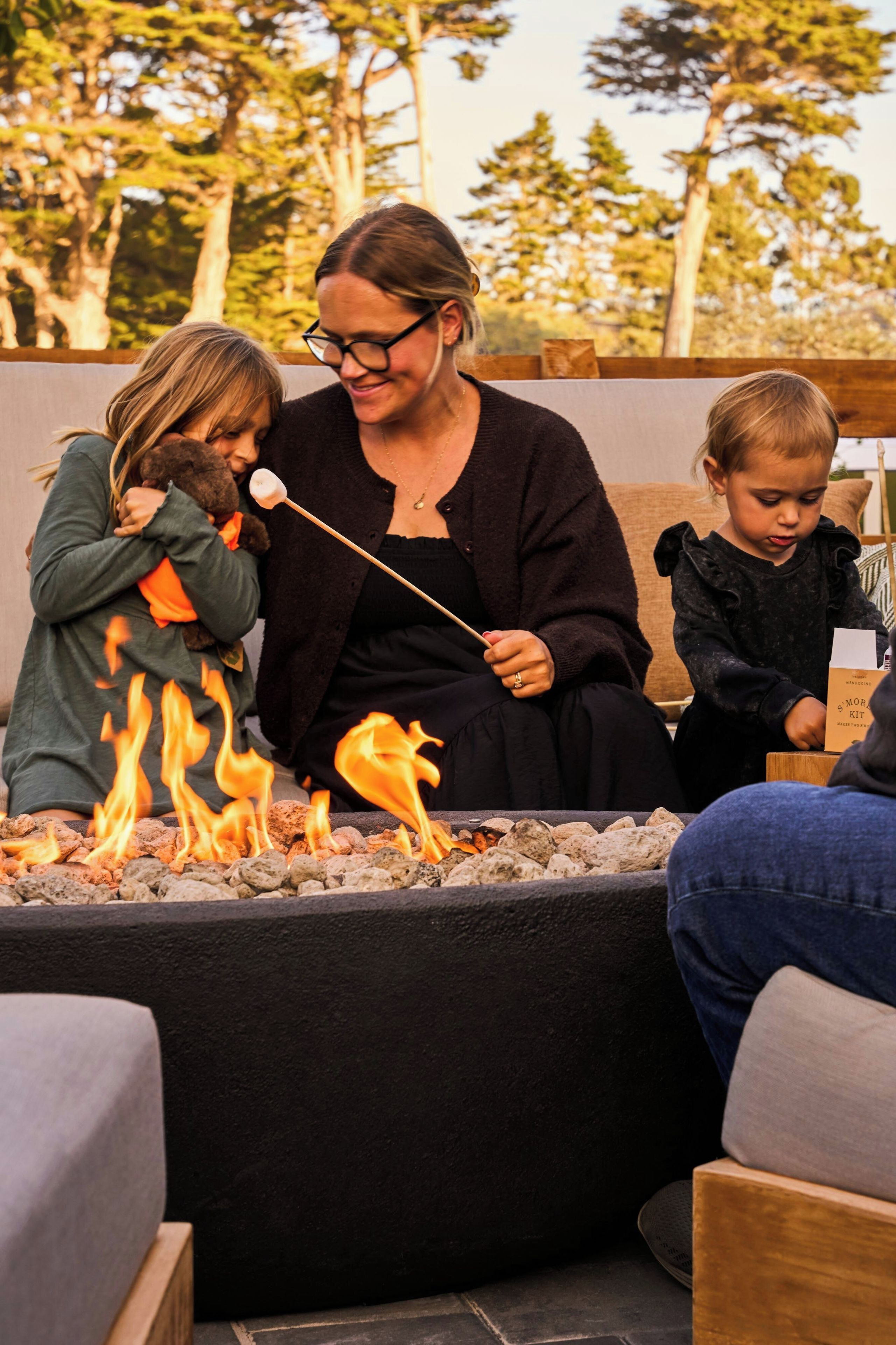 A woman roasting marshmallows over a fire pit with two children.