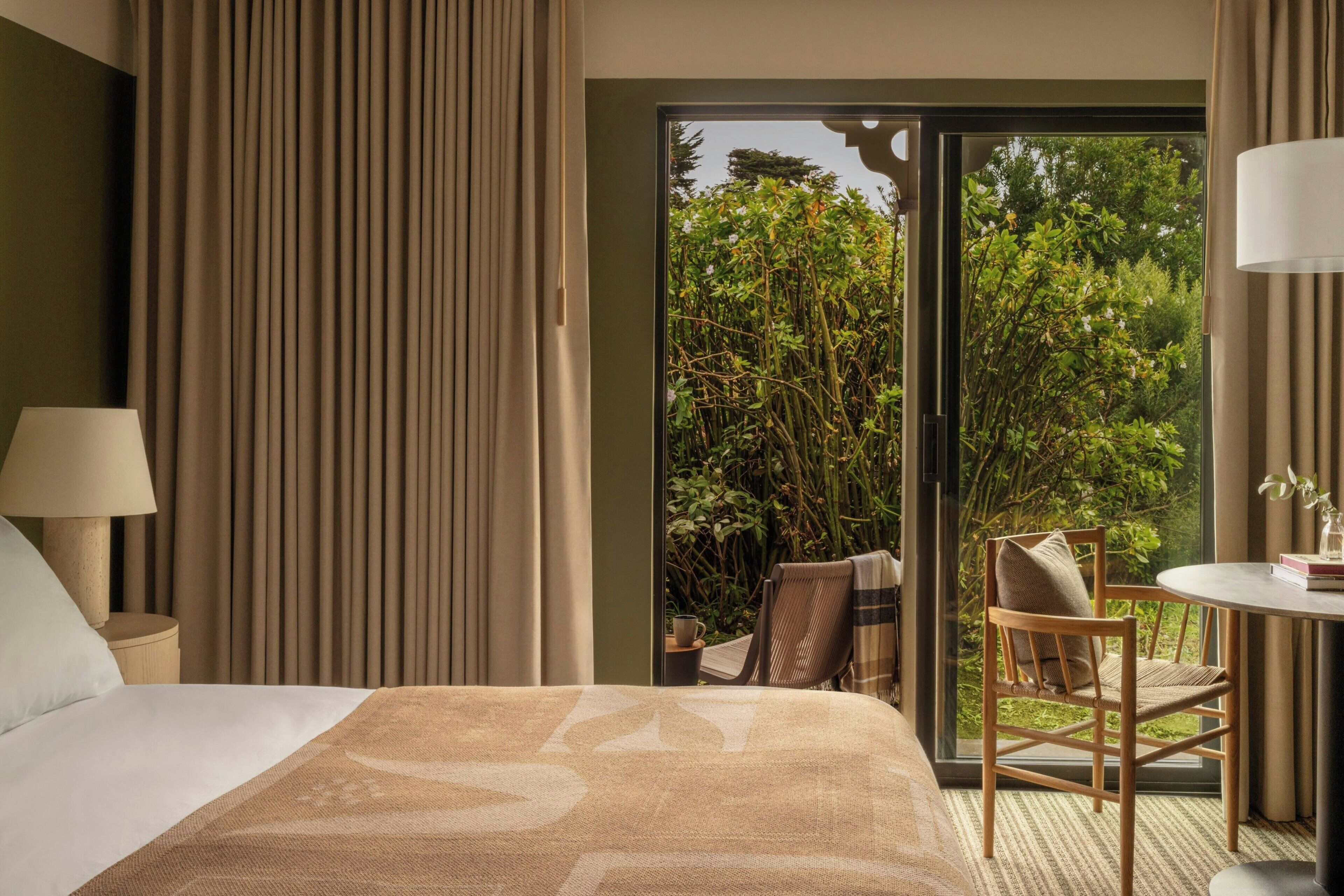 A hotel room with a bed, a chair, a table, and a view of greenery outside.