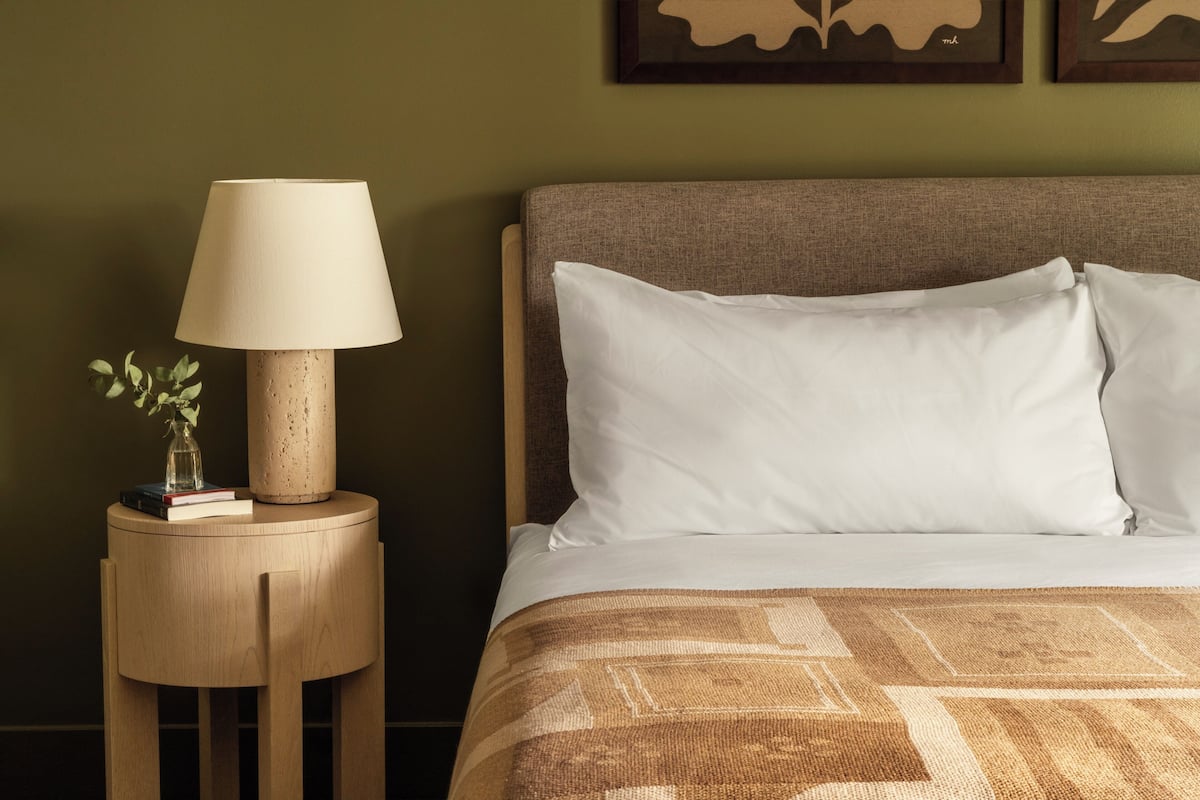 Un lit bien fait, avec des oreillers blancs et une couvre-lit à motifs, une table de chevet ronde en bois avec une lampe et un petit vase de fleurs, et un mur orné d’œuvres d’art encadrées.