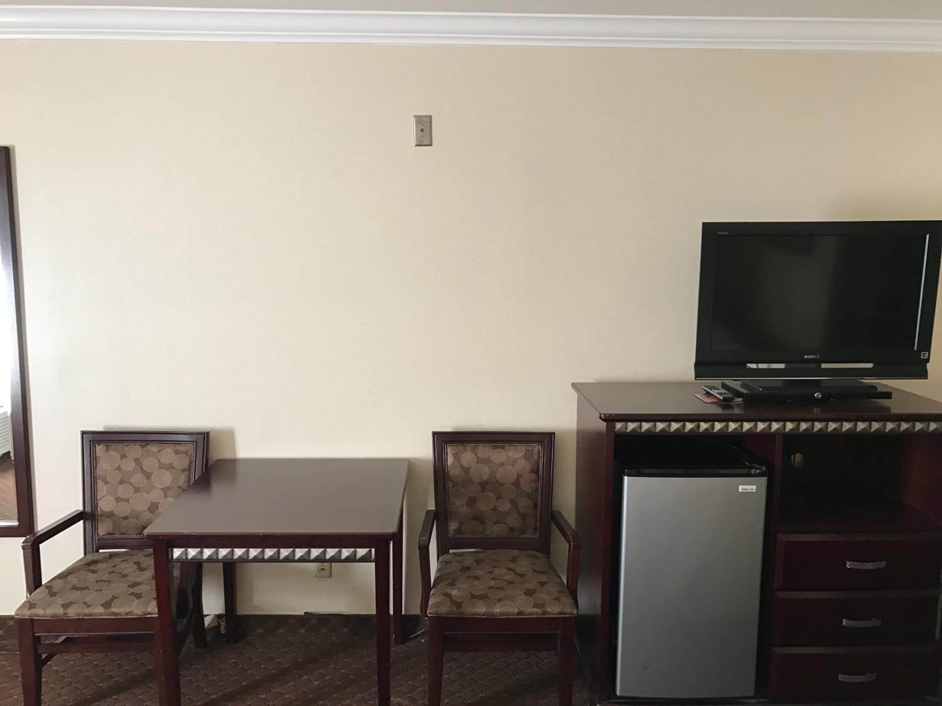 A hotel room with a wooden desk, two patterned chairs, a television, and a mini-fridge.