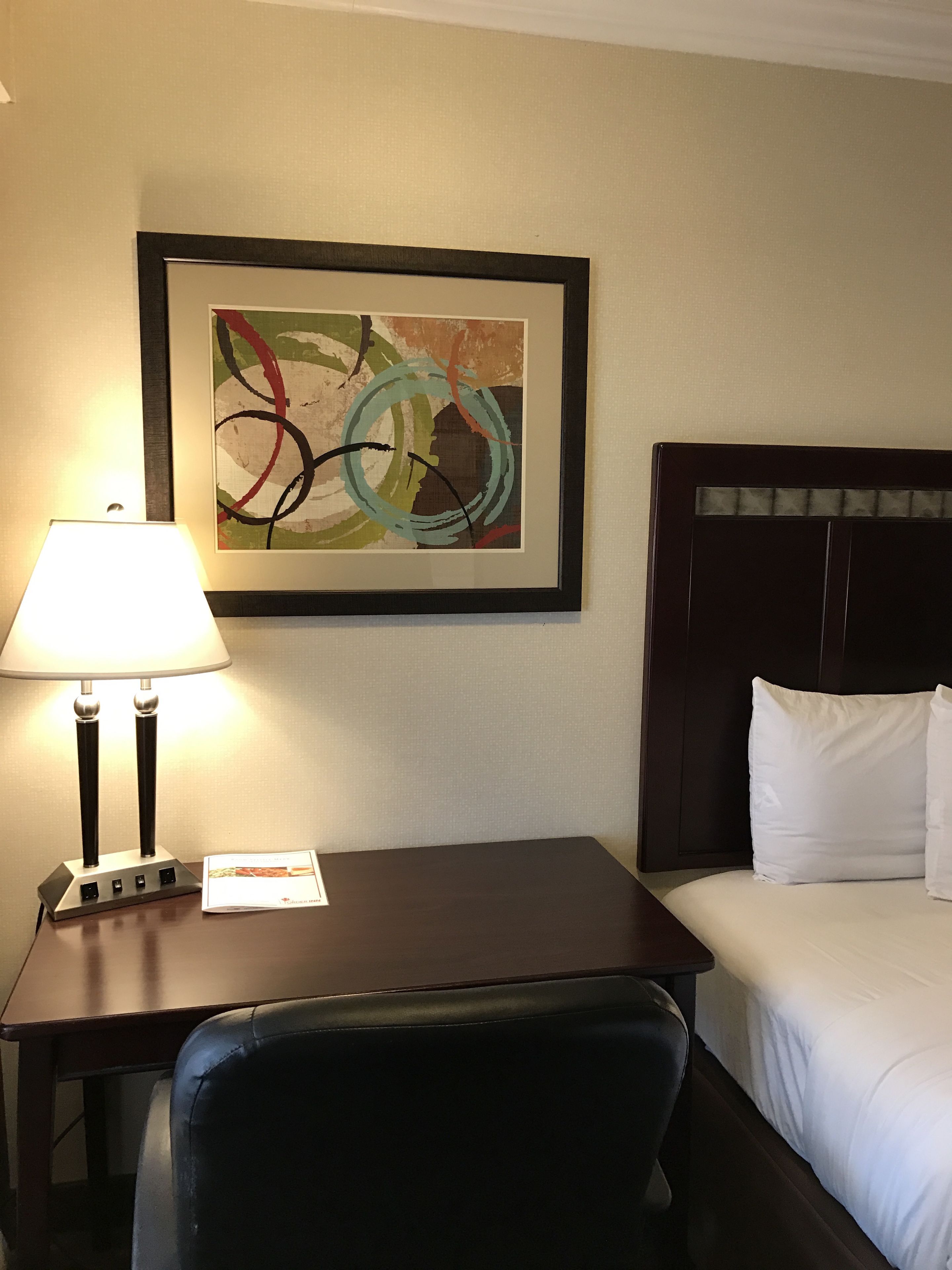 Habitación de hotel con cama, escritorio con lámpara, silla y cuadro abstracto enmarcado en la pared.
