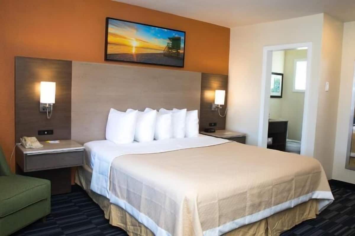A hotel room with a large bed, two bedside tables, a green armchair, a painting on the wall, and a mirror.
